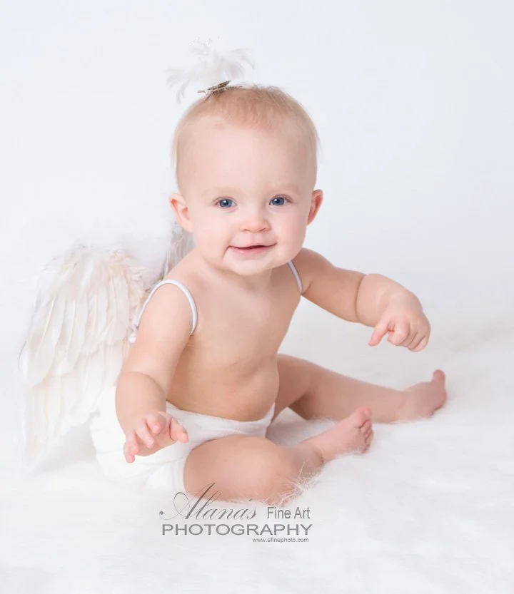 Baby dressed as an angel with wings, sitting on a white fluffy surface.