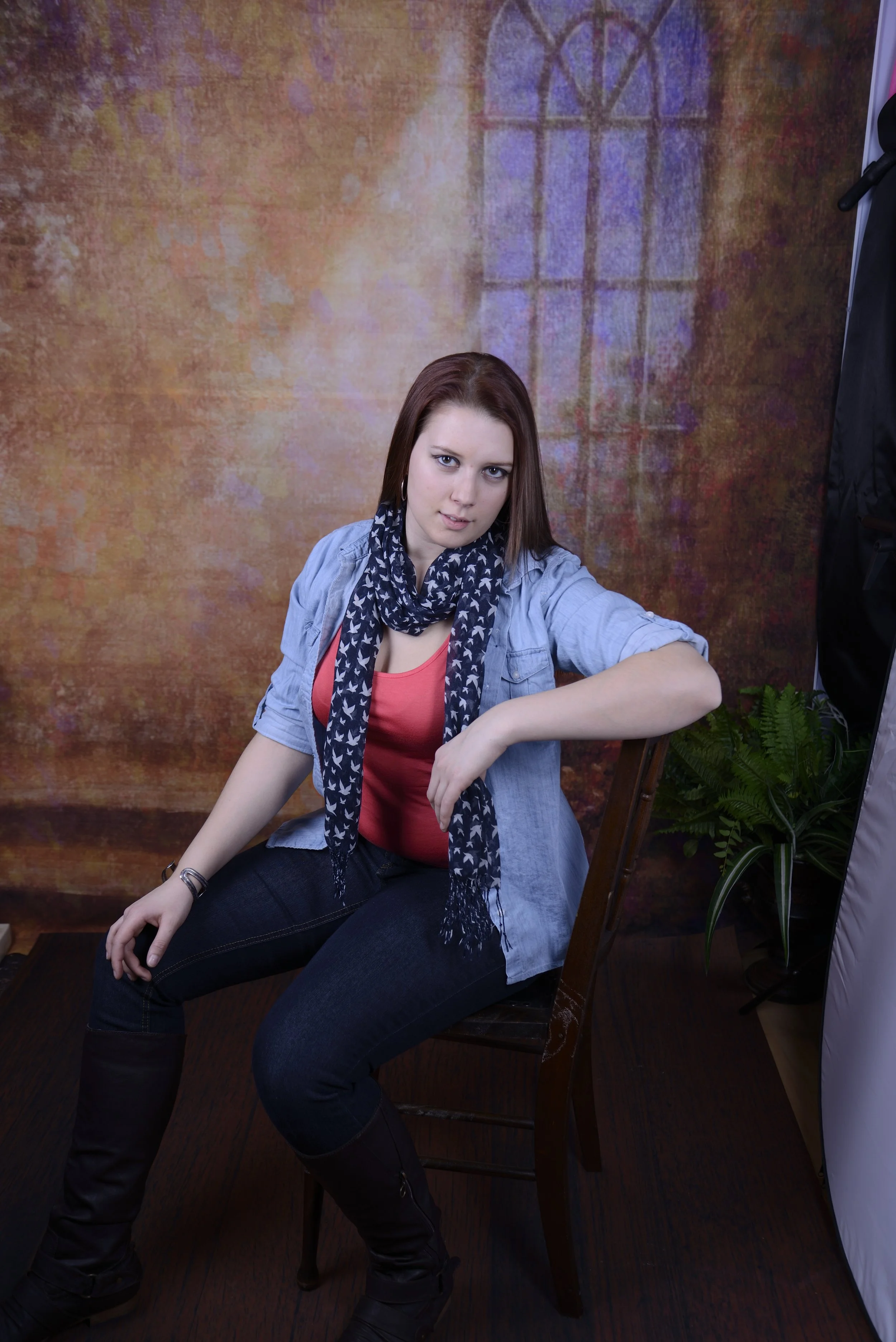A woman sitting on a wooden chair posing with her left arm resting on the backrest, wearing a light denim shirt, pink top, dark jeans, and black boots, with a navy scarf with white patterns around her neck, in front of a textured, colorful backdrop w