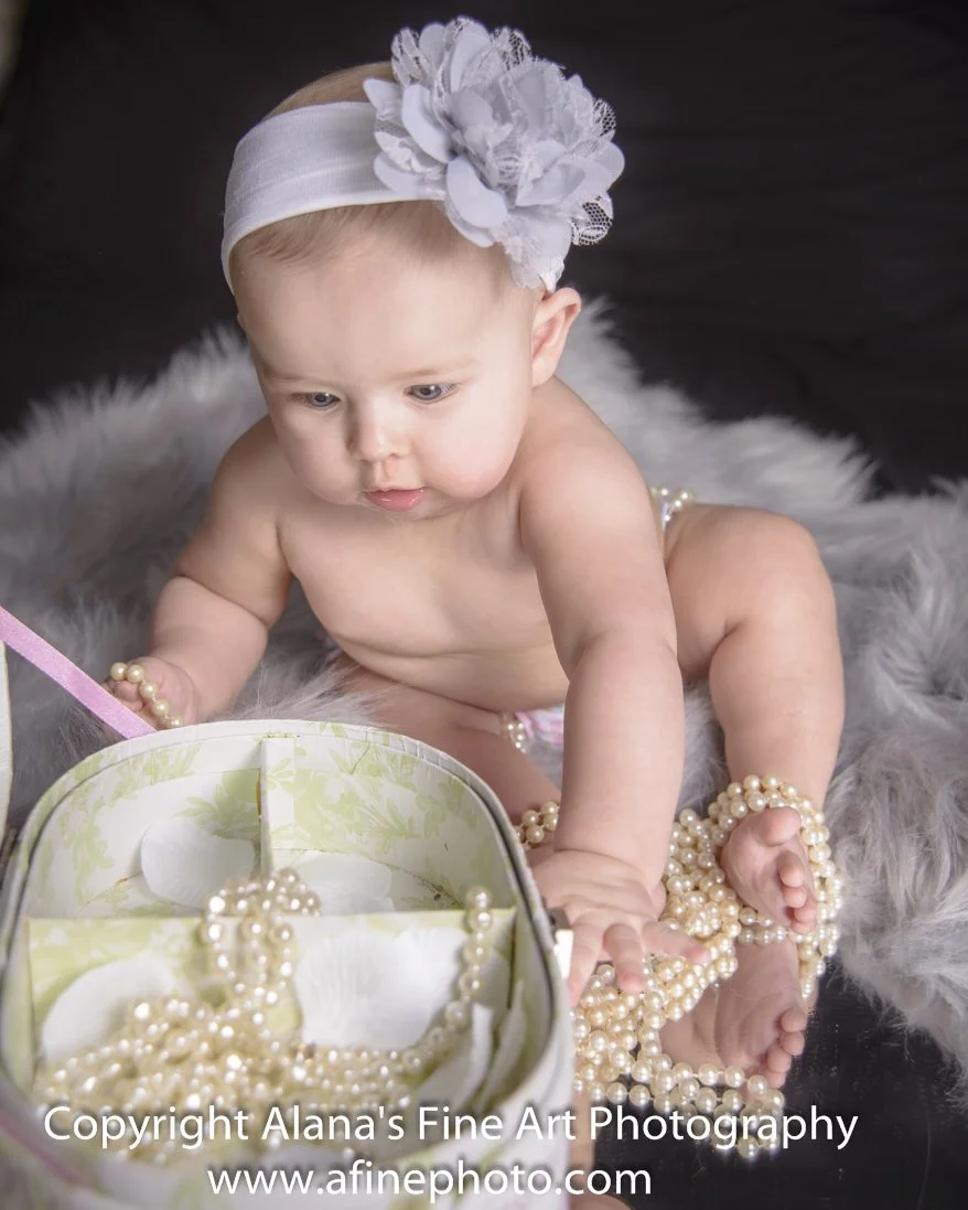 A baby girl sitting on a fluffy gray surface, wearing a white flower headband, exploring a box filled with pearl necklaces.