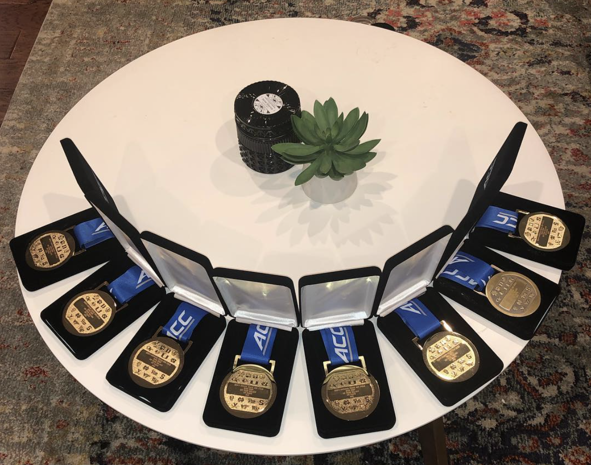 A white round table displays ten gold medals with blue ribbons, arranged in star shape, with three miniature photo frames, a small potted succulent, and a stack of what appears to be black containers or cans in the center, on a multicolored rug.