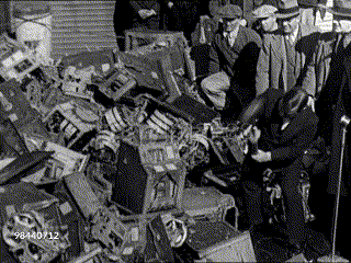 Mayor Fiorello LaGuardia using a hammer to destroy a pile of slot machines observed by several men in suits and hats.