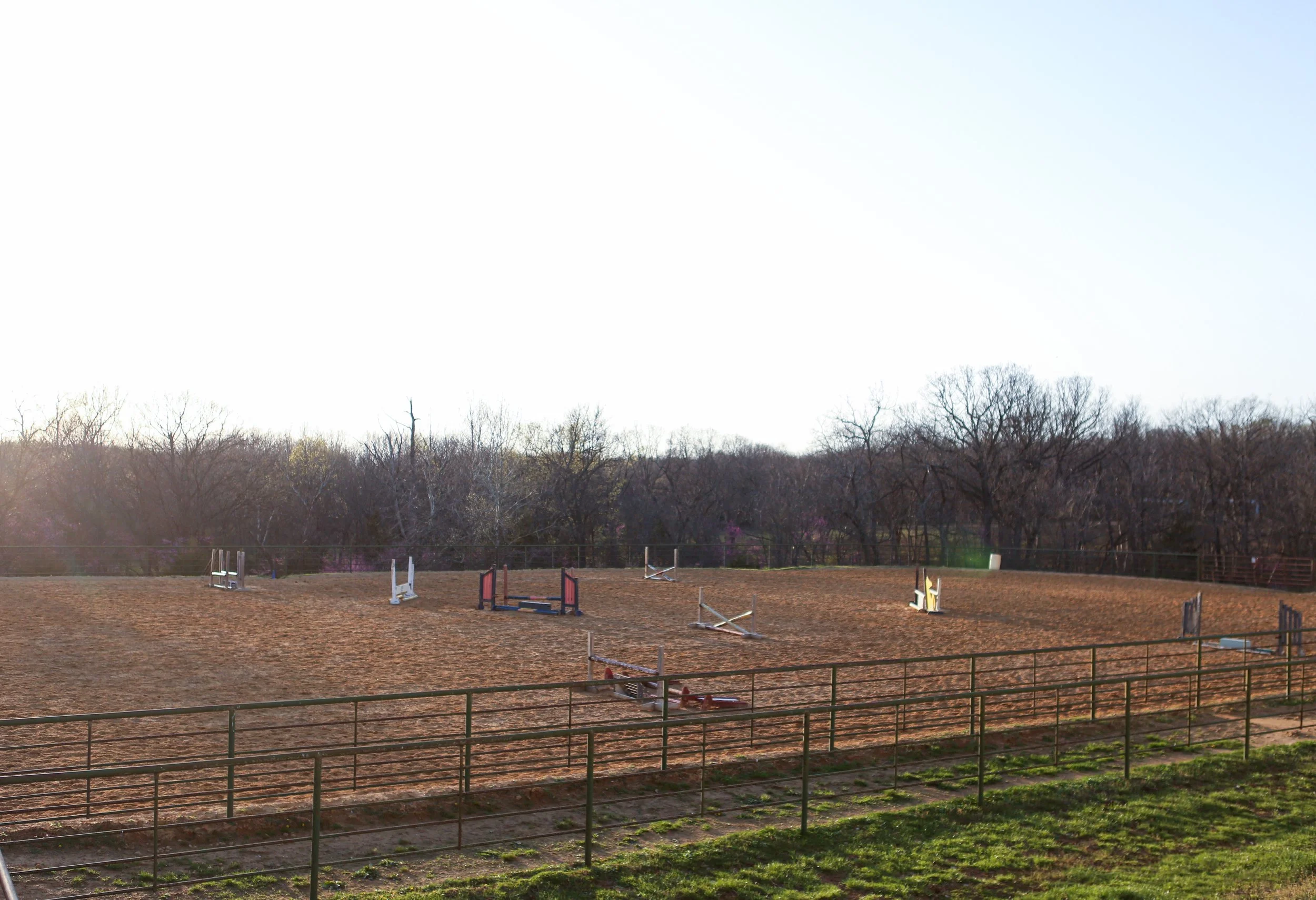An outdoor horse jumping arena with various colorful jumps on a reddish dirt surface, surrounded by a black fence, with leafless trees in the background and a clear sky overhead.