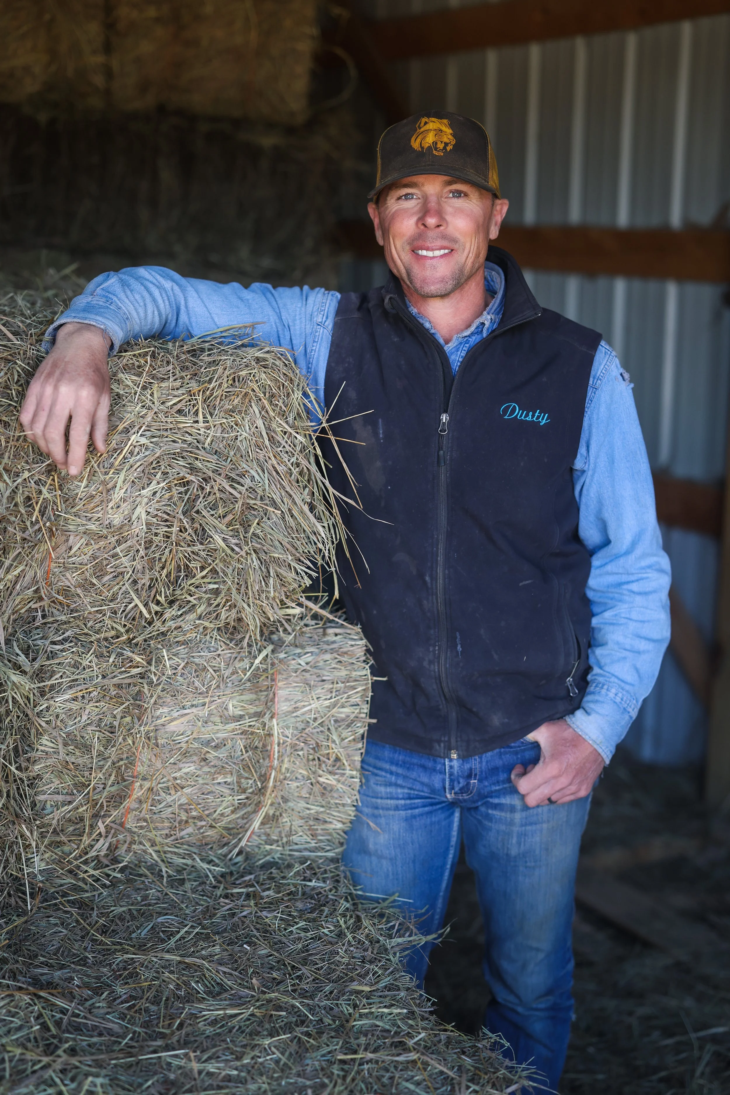 A man in a blue shirt and black vest with 'Dusty' embroidered on it stands next to a round bale of hay inside a barn. He is wearing a baseball cap with a golden lion emblem, smiling and resting his arm on the bale.