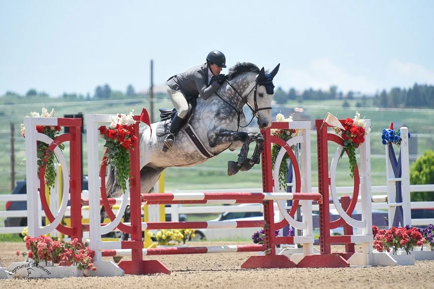 A person riding a horse jumping over an obstacle at an equestrian event, with colorful flowers and decorated fences in a rural outdoor setting.