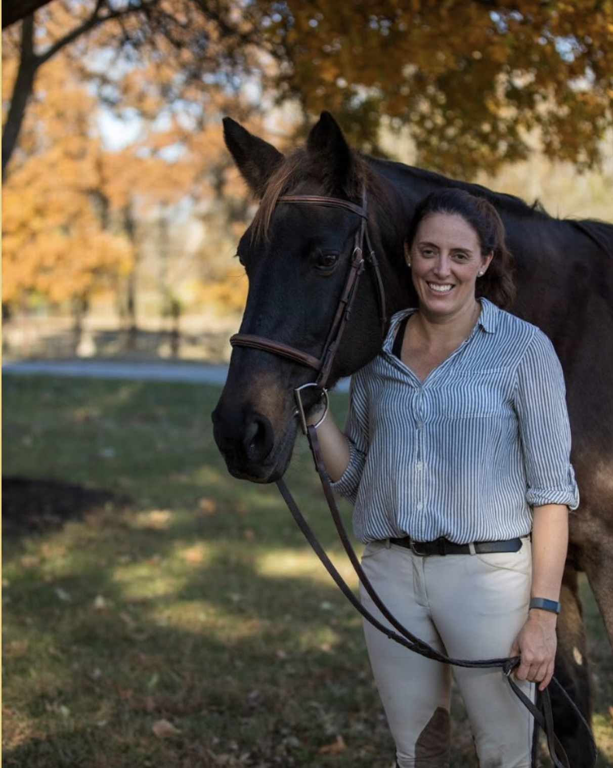 A woman standing outdoors with a black horse, smiling, surrounded by autumn foliage.