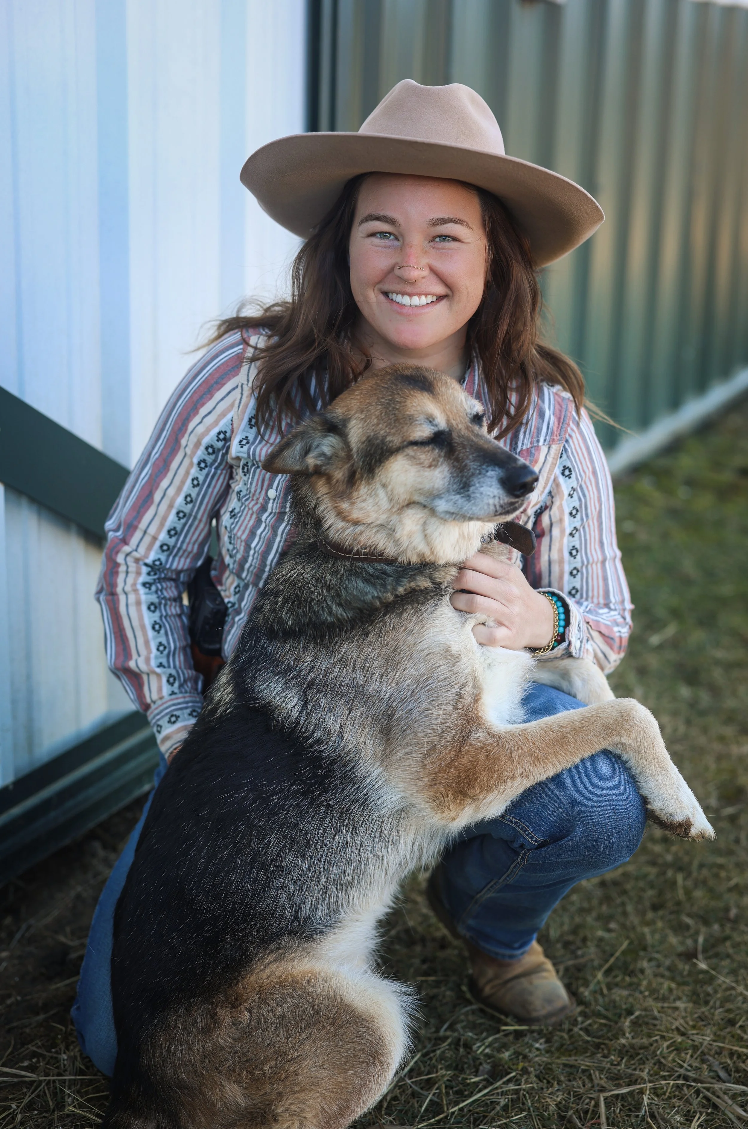 A woman with brown hair, wearing a beige wide-brimmed hat and a striped shirt, smiling while holding a dog with closed eyes and a relaxed expression.
