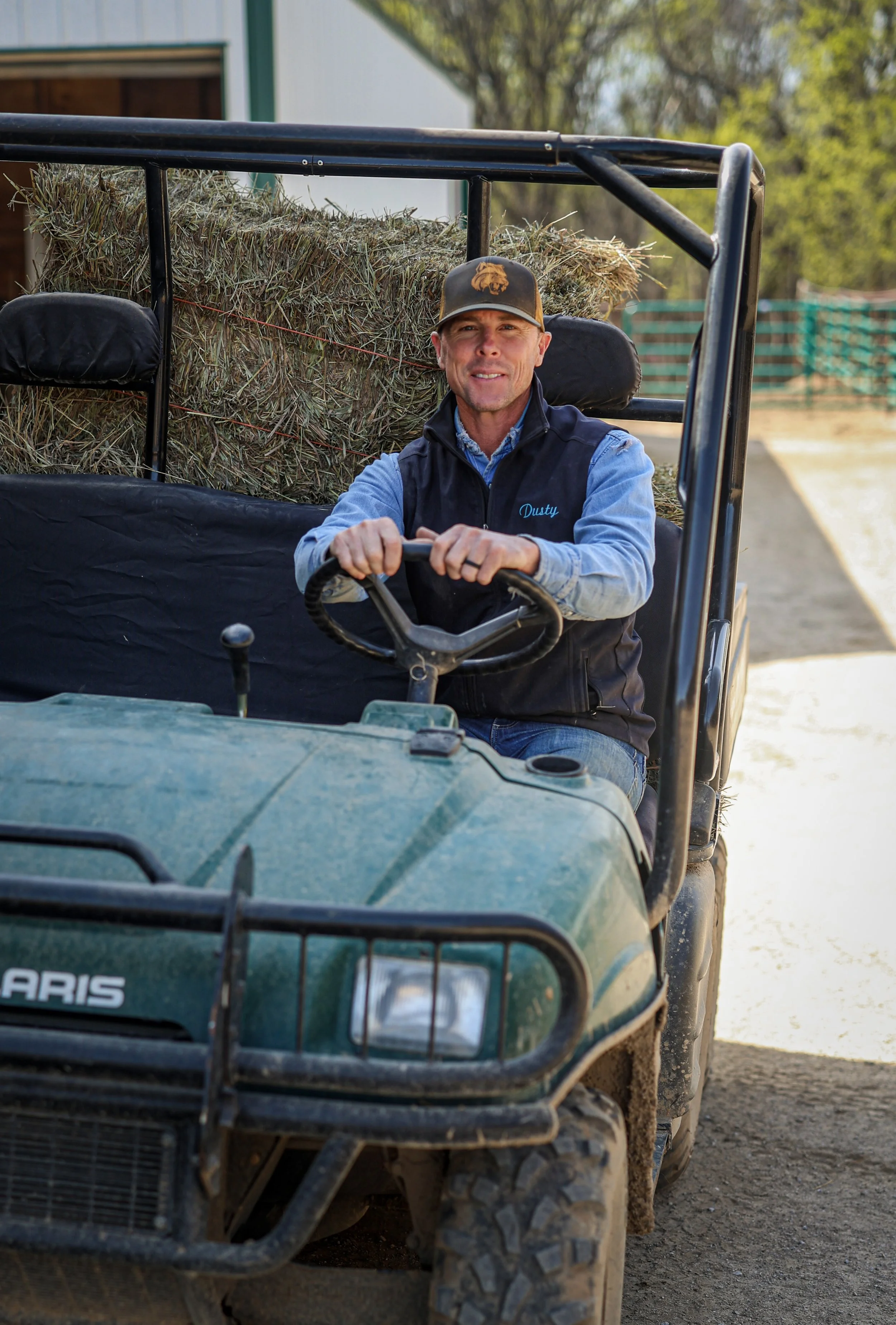 A man wearing a cap and vest sitting on a green Polaris utility vehicle, holding the steering wheel, with a large hay bale behind him.