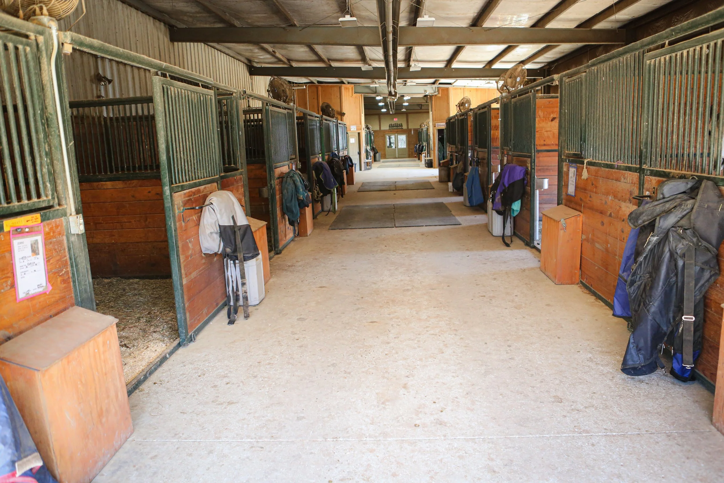 Inside a stable with horse stalls on both sides, some of which have backpacks and jackets hanging on the doors.