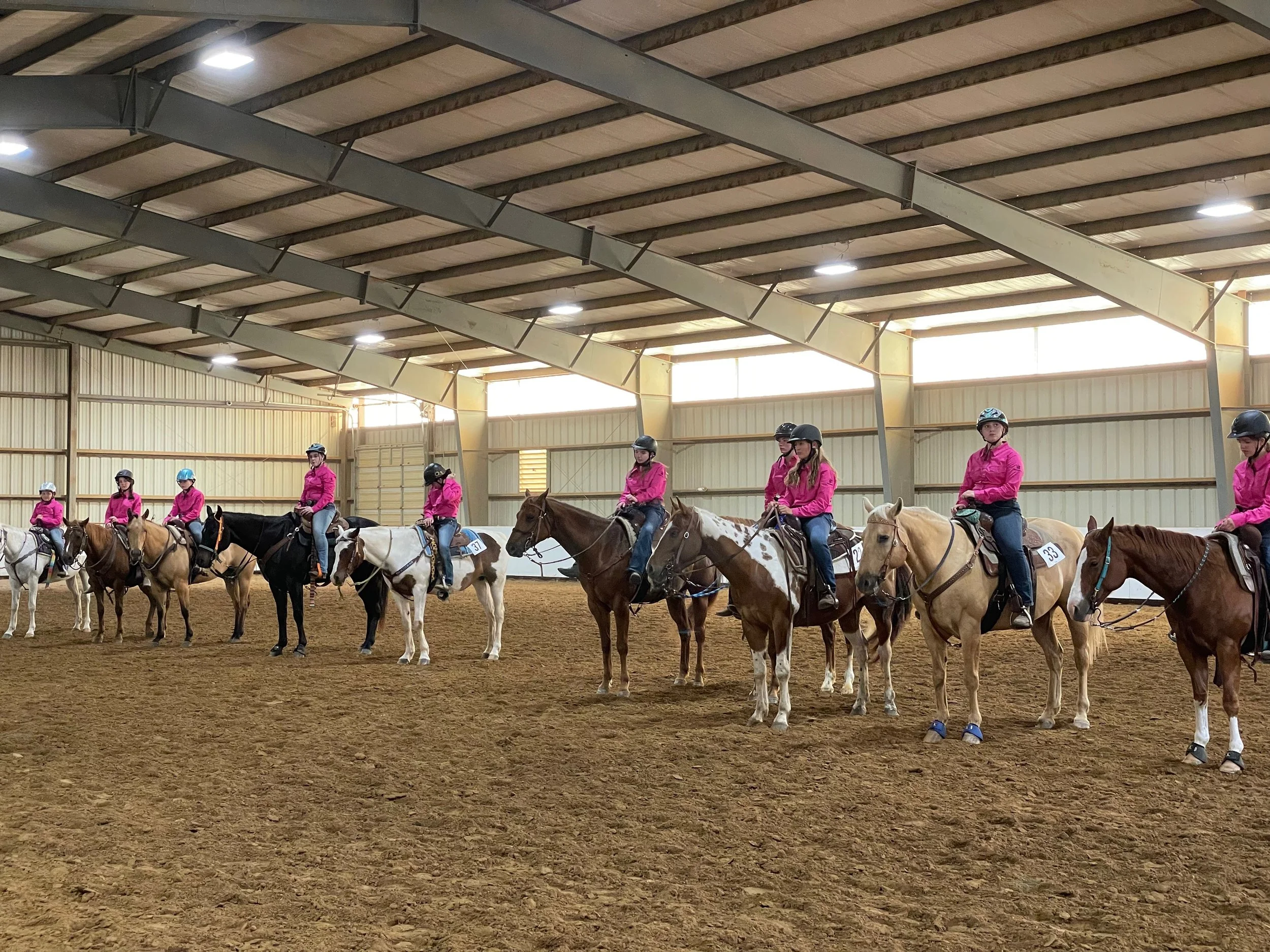 Group of people dressed in pink riding helmets and jackets, mounted on various colored horses, lined up inside a spacious indoor riding arena with a dirt floor and metal walls.