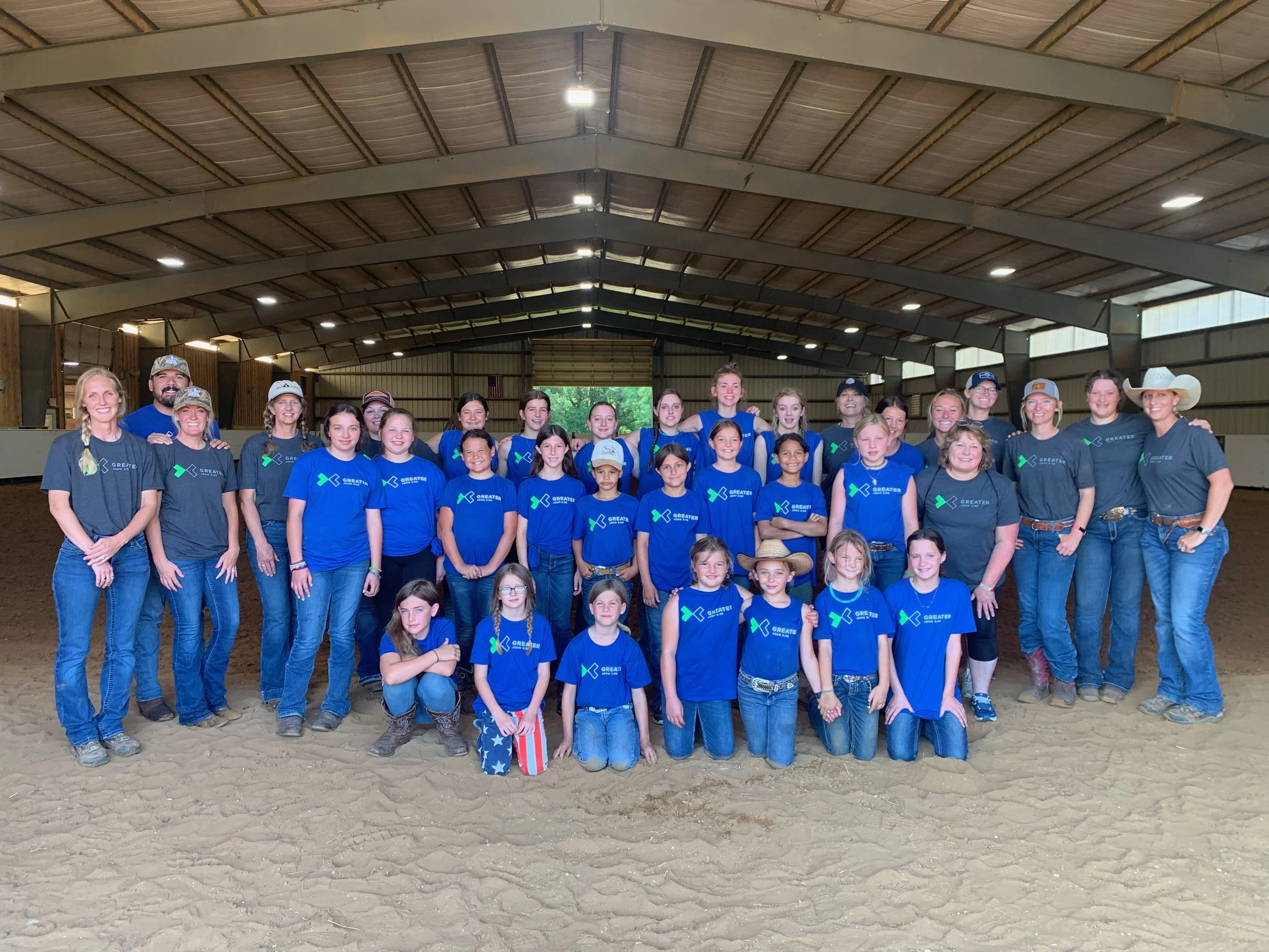 Group photo of children and adults in an indoor riding arena, some wearing Blue and some wearing Gray shirts, all smiling and posing for the camera.
