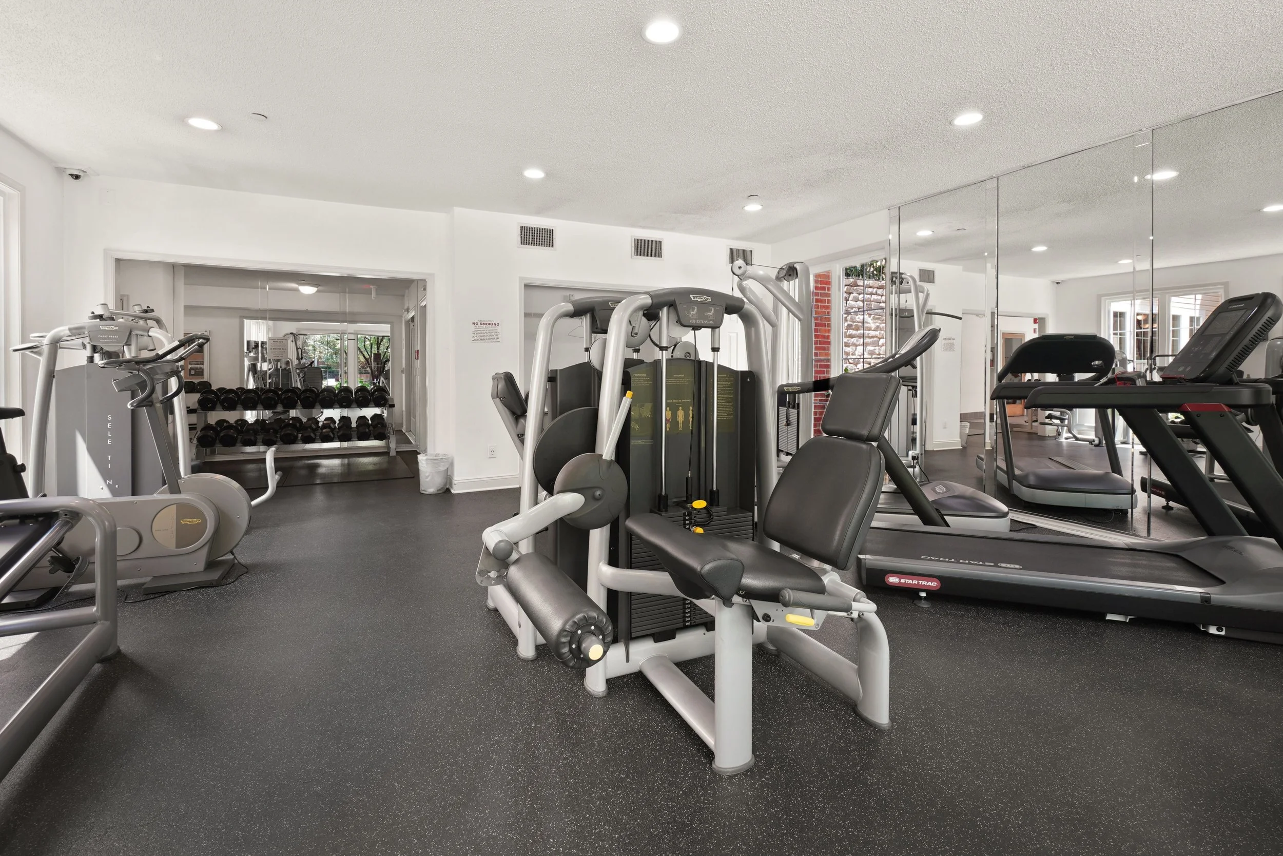 A modern gym room with various exercise equipment including stationary bikes, weight machines, and treadmills. Large mirrors cover one wall, and there is a window providing natural light.