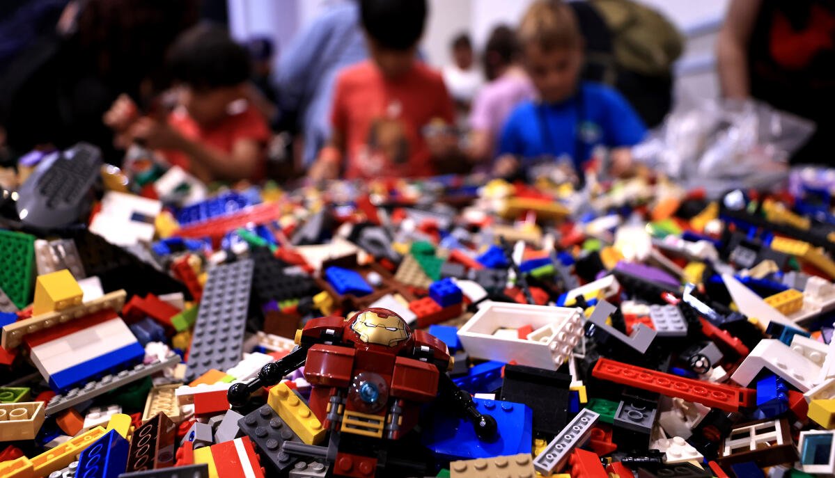 A large pile of assorted LEGO bricks and pieces with a small Iron Man LEGO figure on top. In the background, children are looking at the Lego collection at a crowded event, Brick Blast Live, LEGO Convention