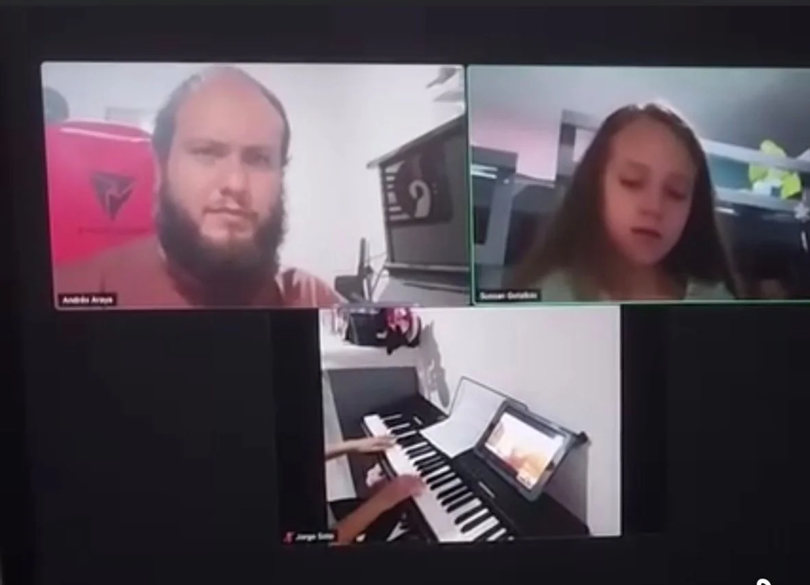 Videollamada con tres personas, dos en la parte superior y una pantalla compartida que muestra manos tocando un teclado musical. Las personas se ven en ventanas separadas en una plataforma de videoconferencia.
