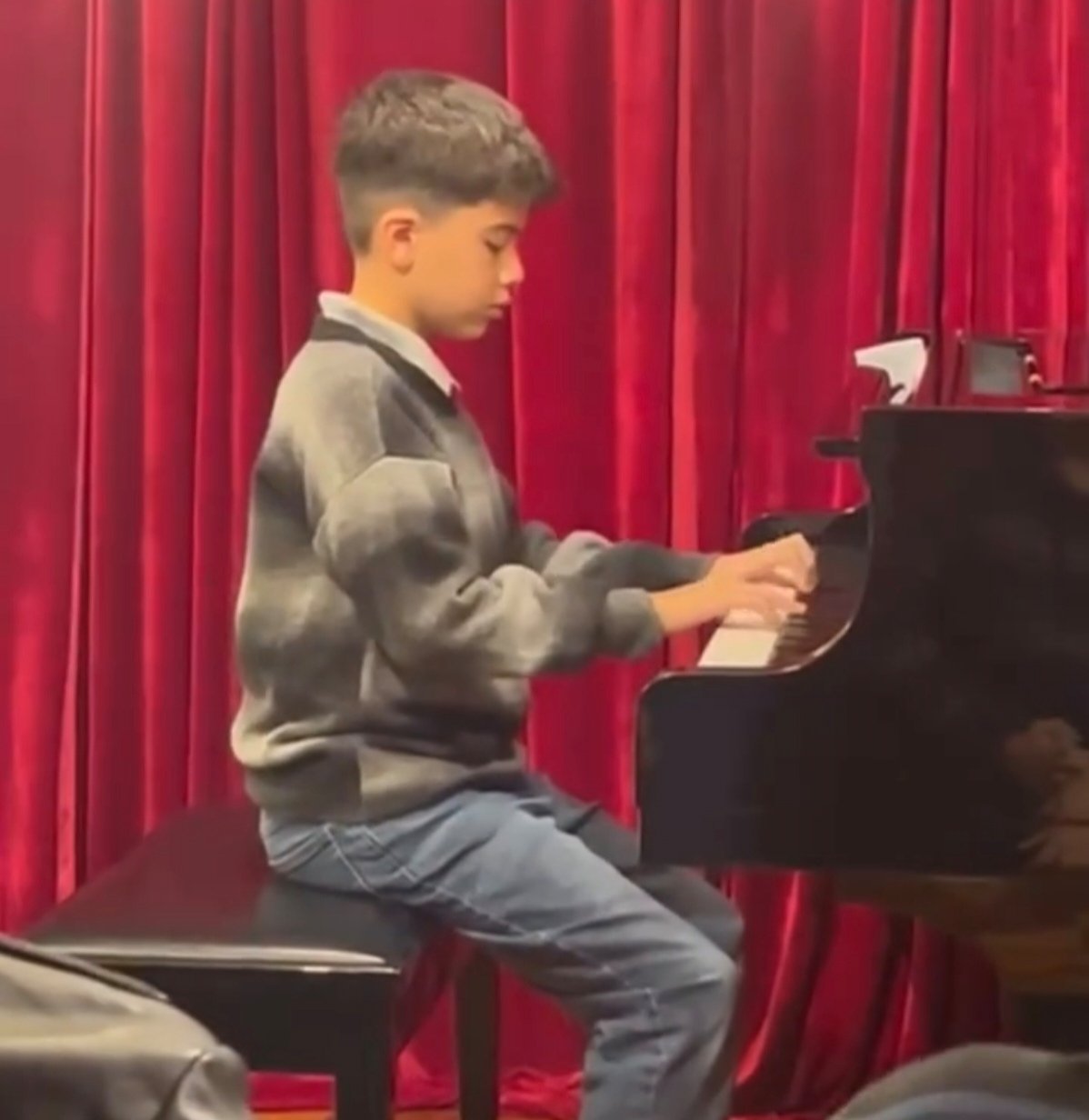 Niño tocando el piano frente a una cortina roja, concentrado en la música.