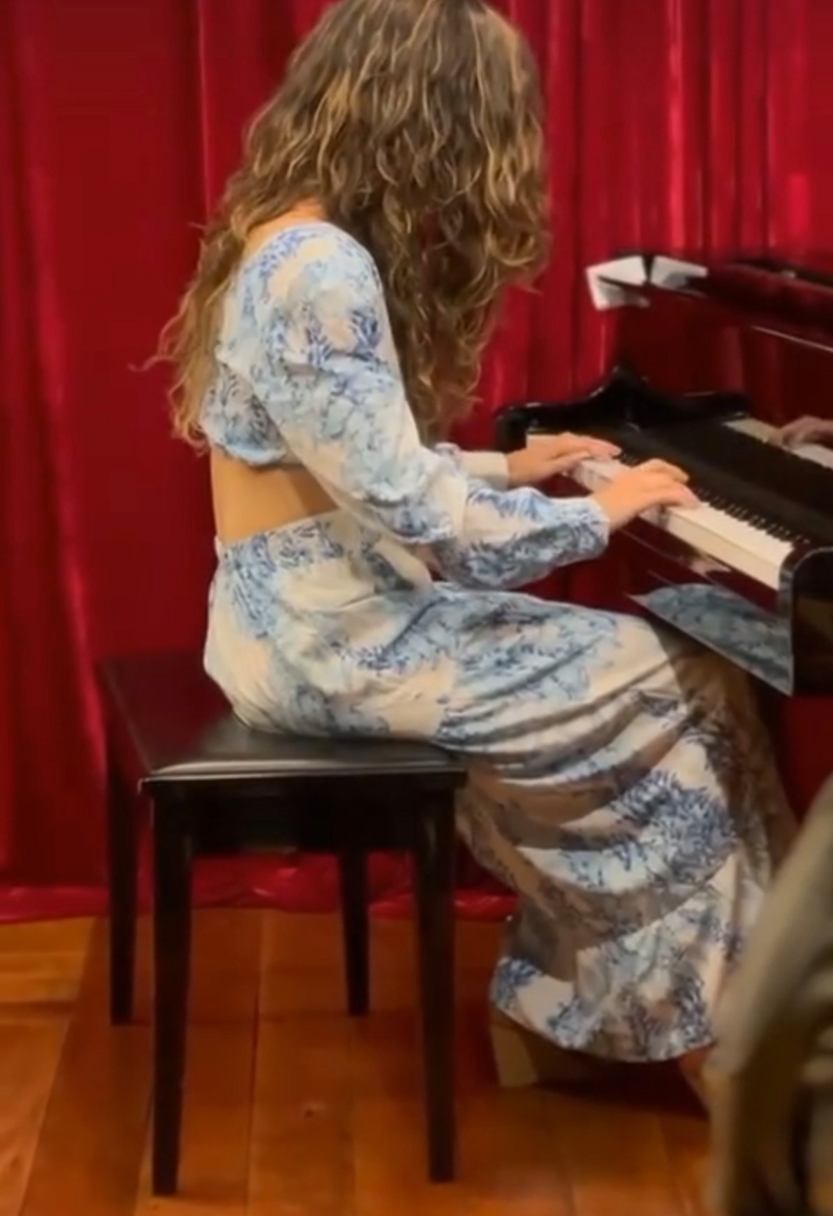 Persona tocando el piano con vestido azul y blanco, cortinas rojas de fondo.