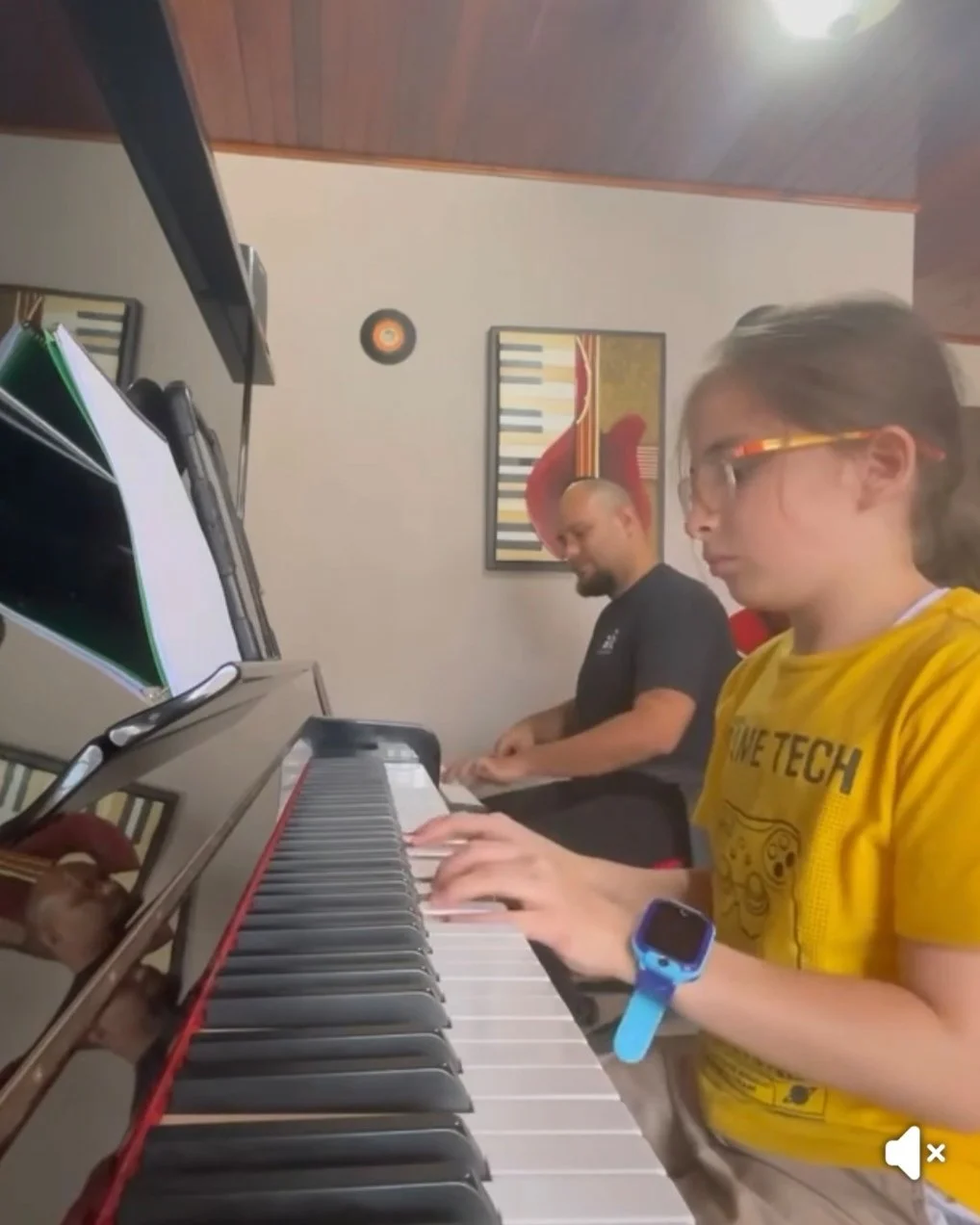 Niño tocando el piano con un hombre al fondo, en una sala con decoración musical.