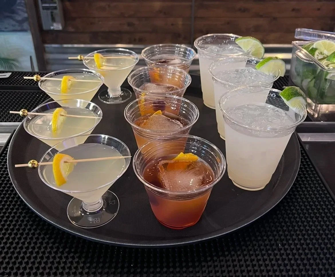 Arrangement of cocktails and drinks on a black tray, including margaritas with lemon wedges, iced tea with lemon slices, and clear drinks with lime wedges, on a bar or outdoor setting.