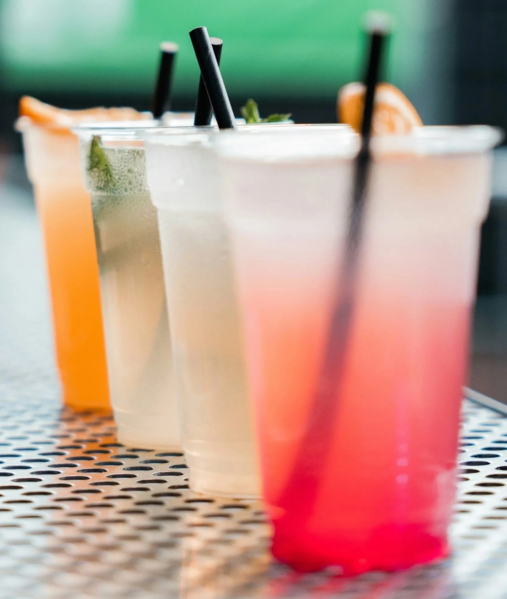 Four colorful beverages with straws on a perforated metal table, with a green and blurred background.
