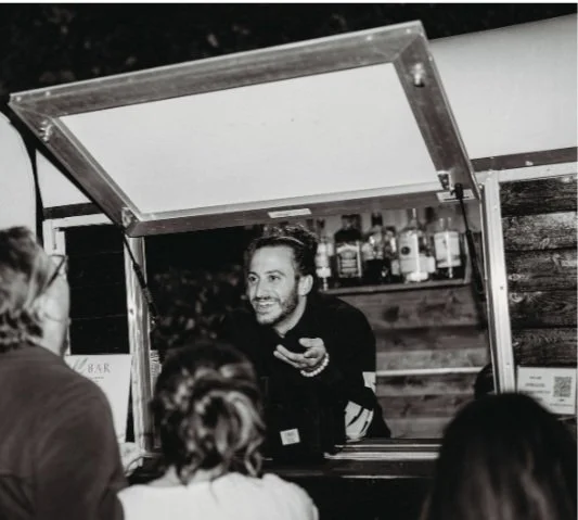 A man inside a horse trailer Mobile bar serving customers outdoors at night.