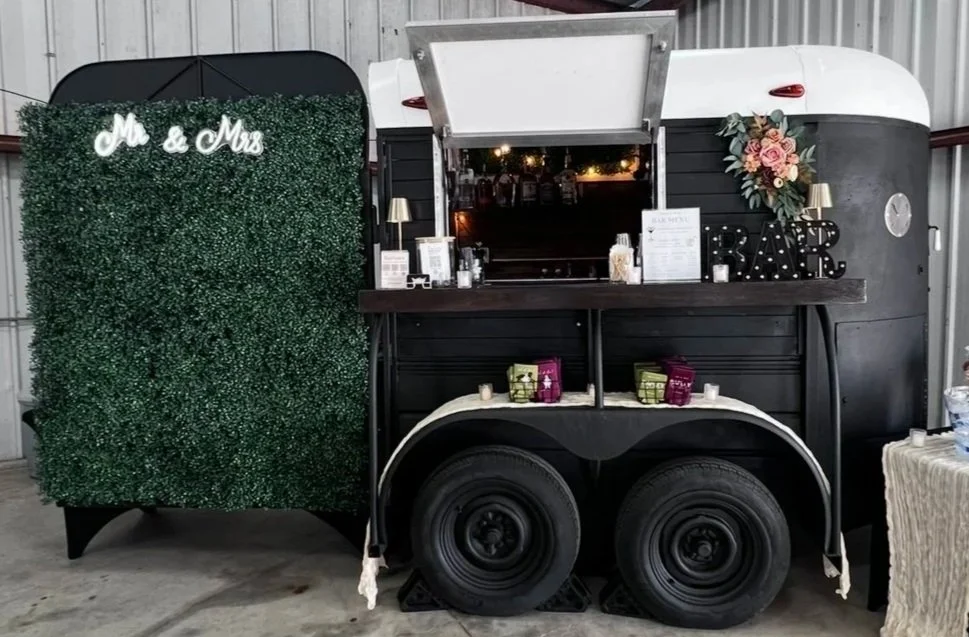 Black Mobile bar horse trailer with a floral decoration and a green hedge wall with 'Mr & Mrs' sign, serving as a bar at a wedding reception.