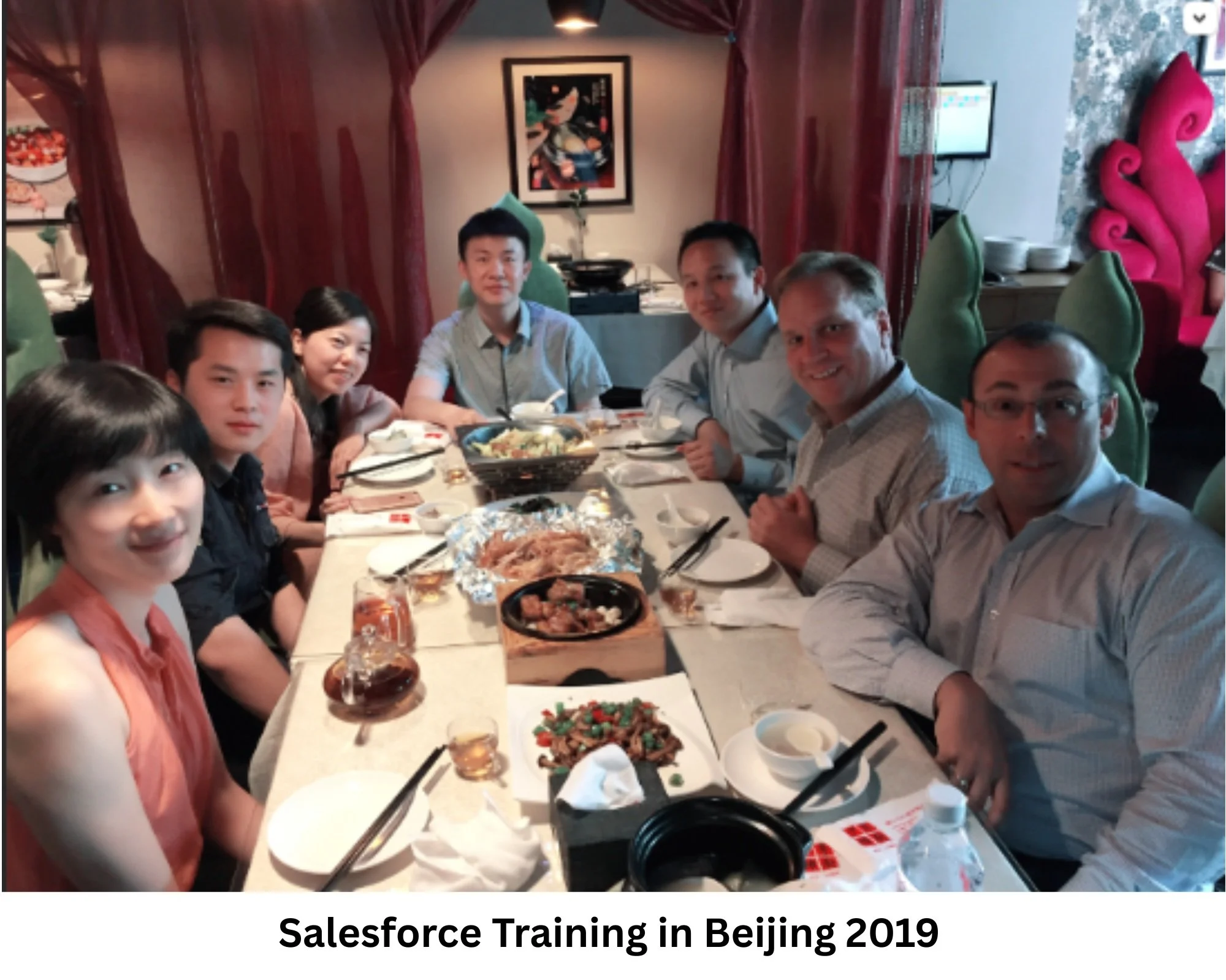 A group of people sitting around a banquet table with food, in a restaurant setting. There are seven individuals, some smiling, with dishes like vegetables, meat, and drinks on the table. The background has Asian-inspired decor and artwork.