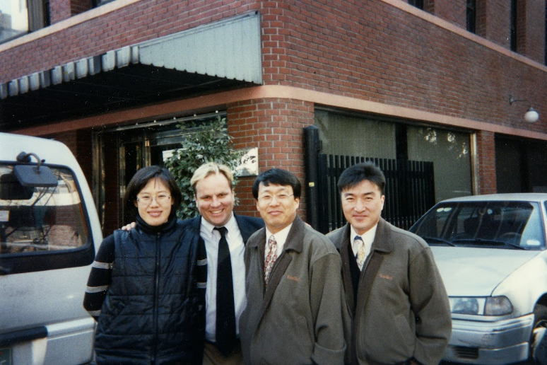 Four people standing together outdoors in front of a building and vehicles, smiling at the camera.