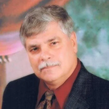 A middle-aged man with gray hair and a mustache posing for a portrait. He is wearing a dark blazer, a maroon shirt, and a patterned tie, with a colorful blurred background.