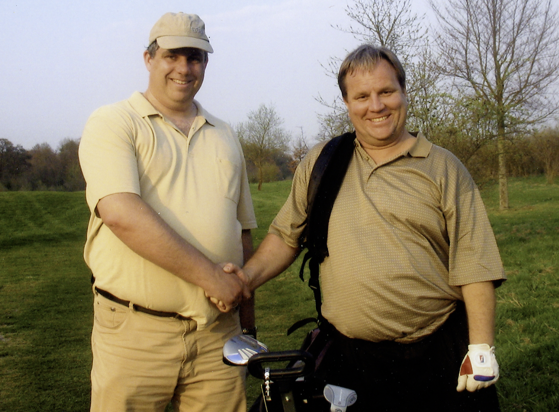 Two men shaking hands on a golf course, smiling at the camera, one with a golf club, and there are trees in the background.