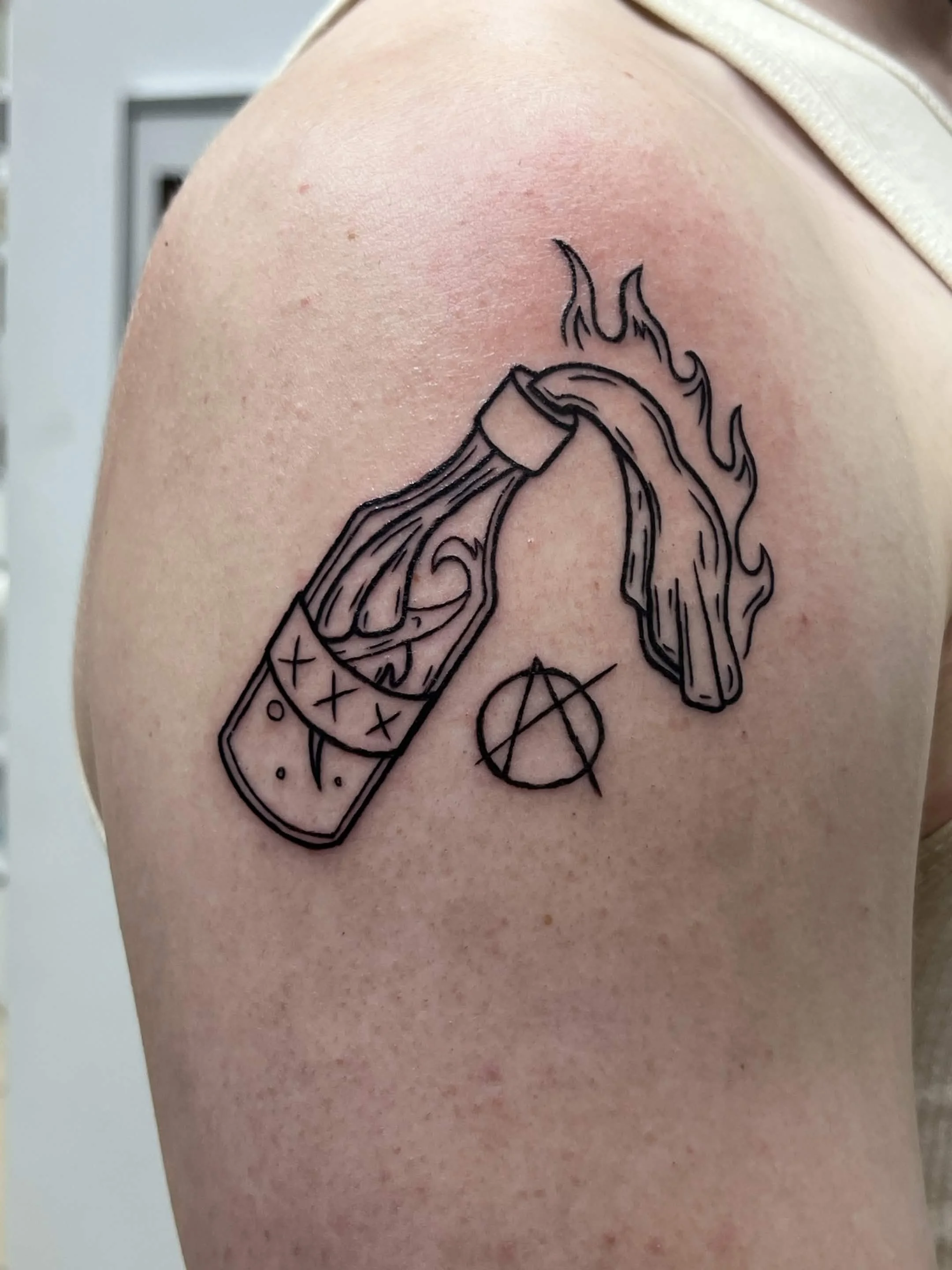 Black line tattoo of a hand holding a stick of dynamite with flames at the top, and symbols with a circle and X below, on a person's upper arm.