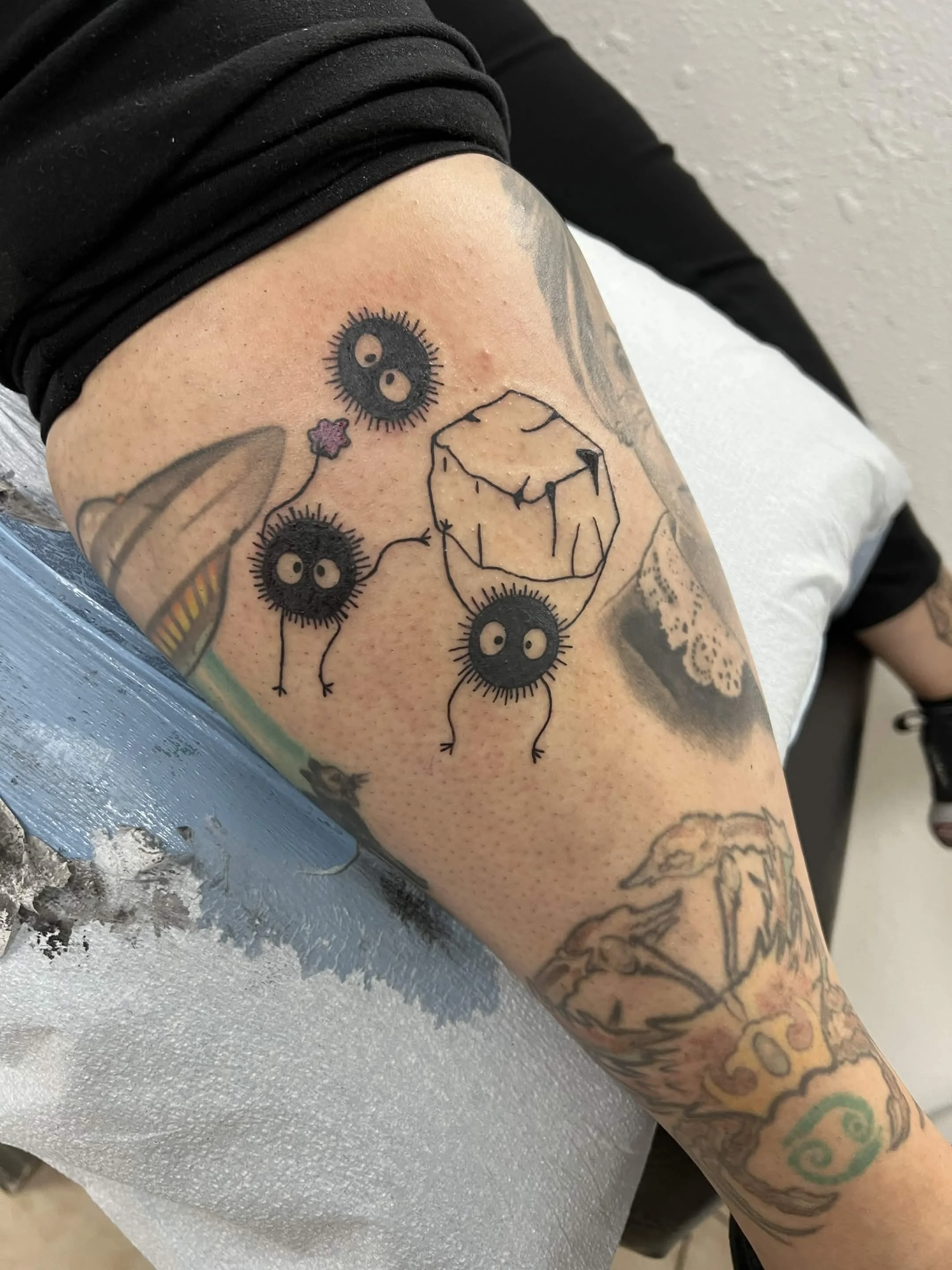 A person's arm with tattoos, including a design of black spiky circle faces with white eyes, a simple outline of a cube, and other tattoo artwork. The person is wearing a black long-sleeve shirt pulled up to expose the tattoos.