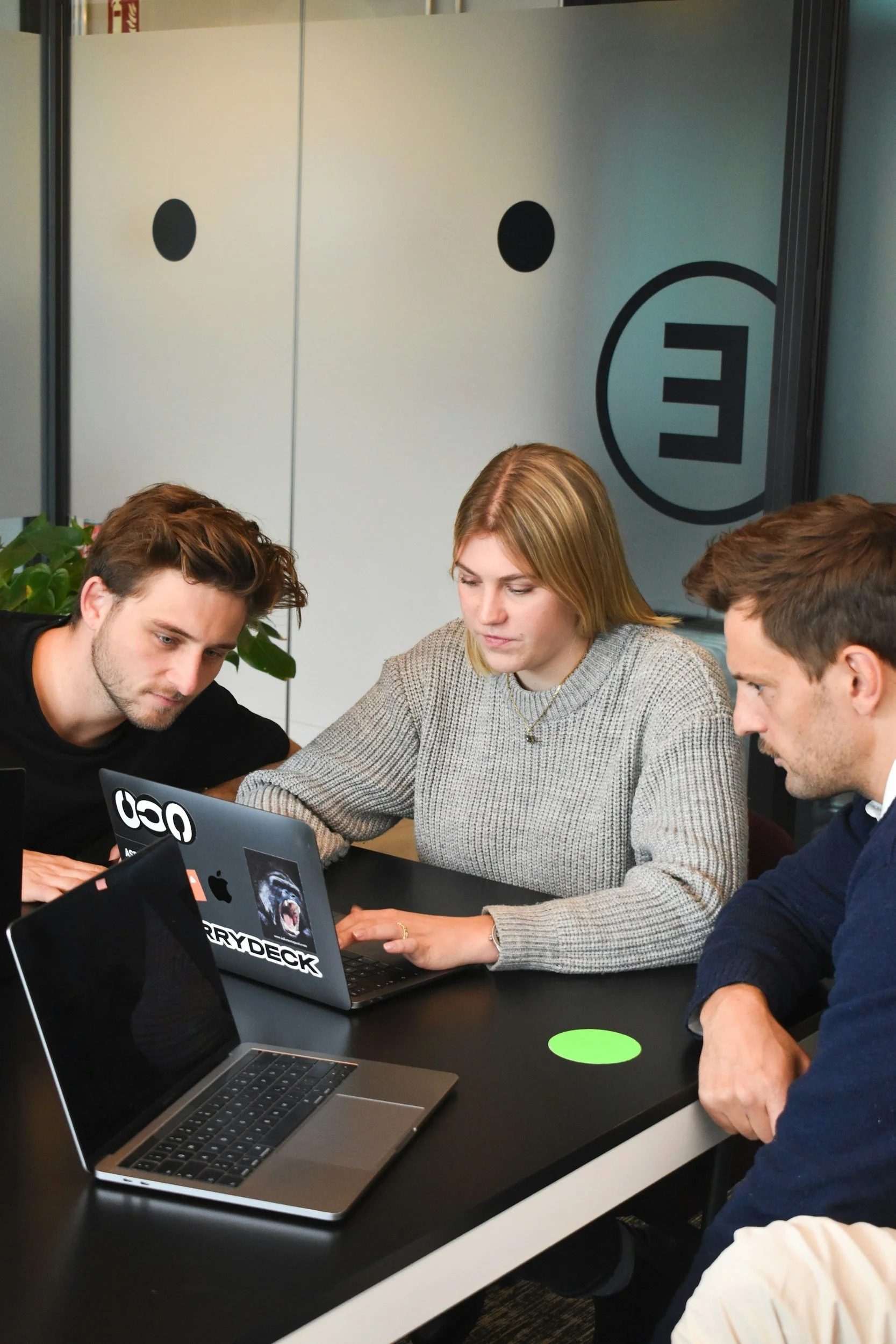 A group of four people sitting at a black conference table focusing on their work with laptops in an office setting. There are two men and a woman close together, with a partial view of another person on the right. The background features glass panels with black circular designs and a large letter 'E' in a circle.