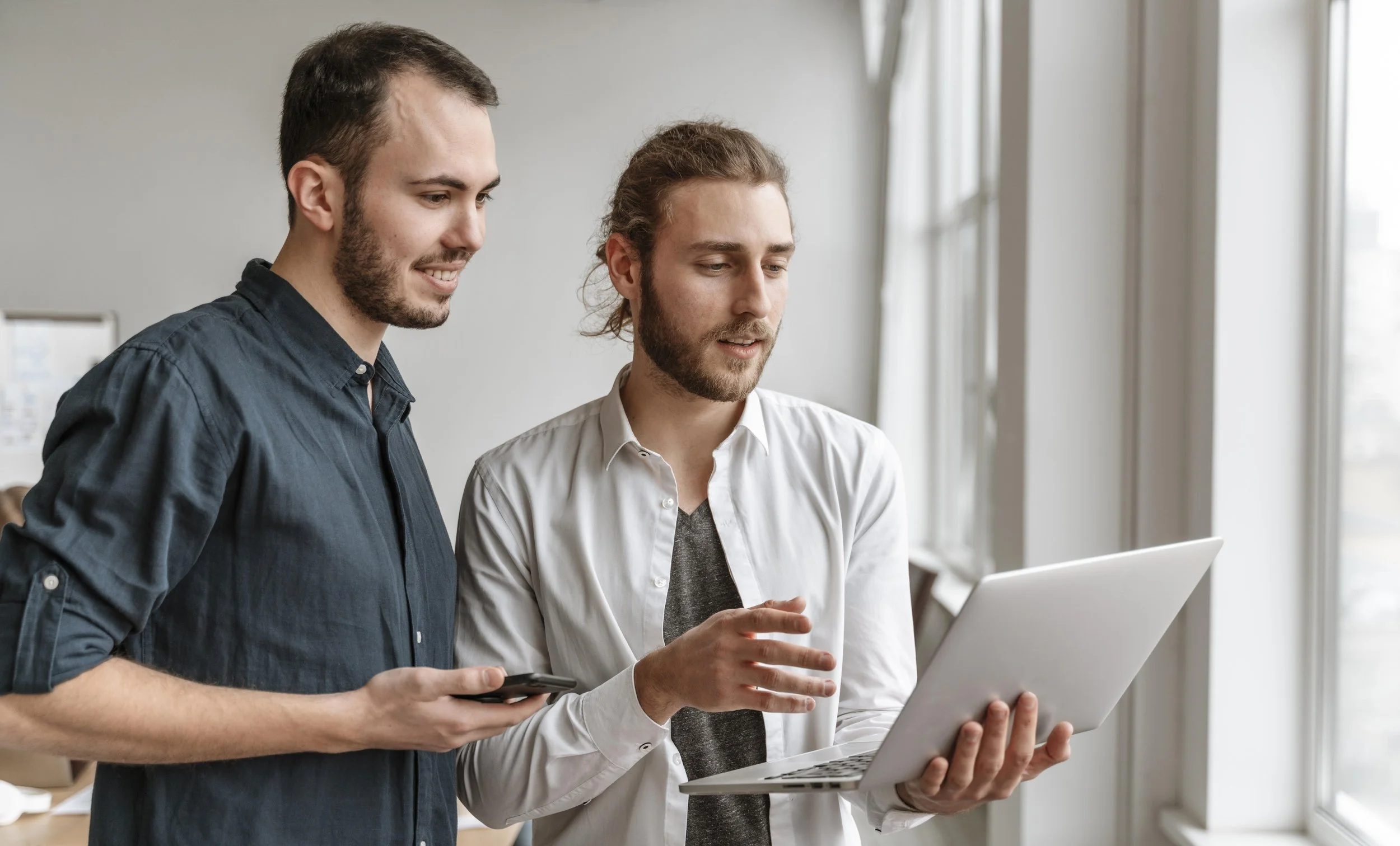Two men engaged in a discussion while looking at a laptop screen, standing by a window in a bright modern office.
