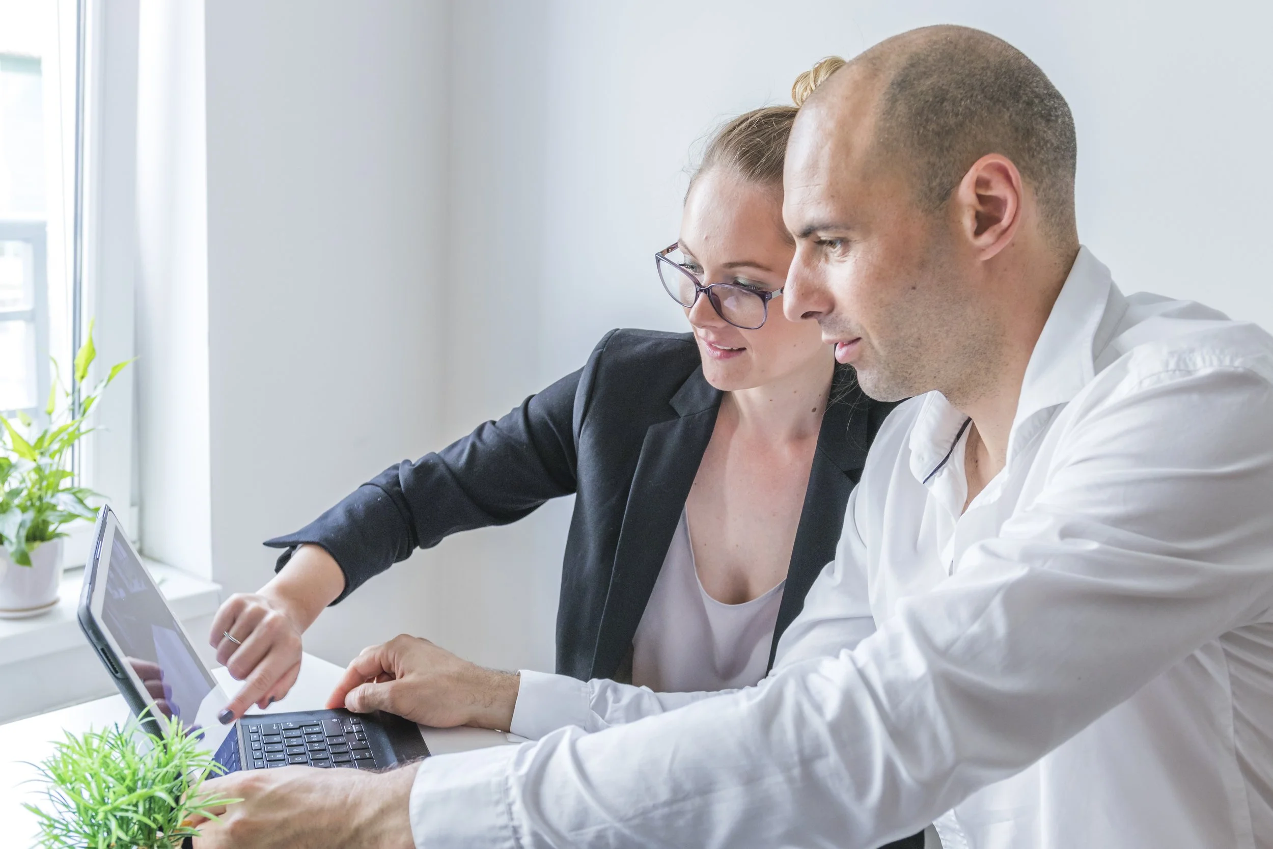 Two professionals, a woman with glasses and a man, working together on a laptop in a bright office. Potted plants are visible on the desk.