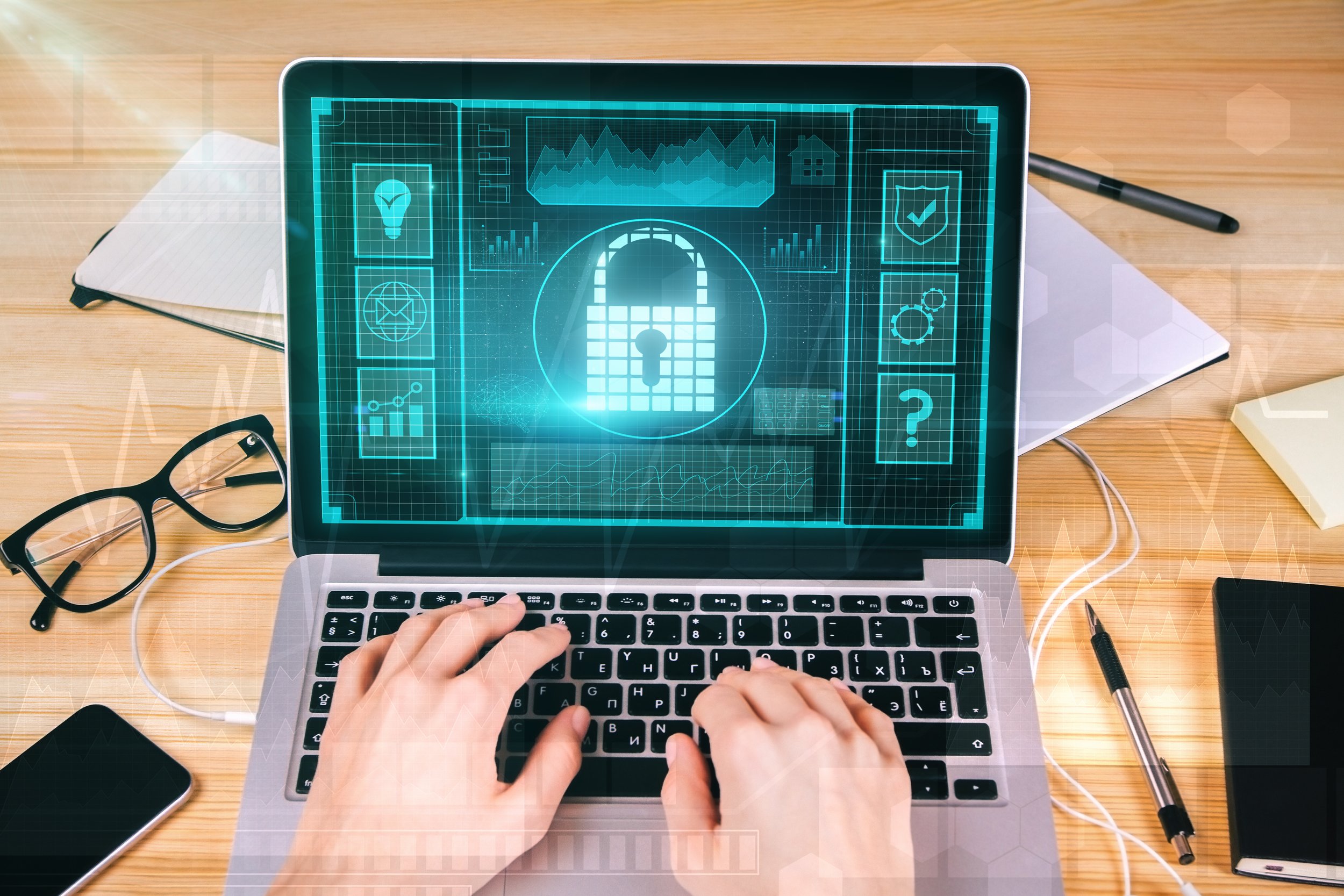 A person typing on a laptop with a digital security interface overlay, showing a padlock and security icons, on a wooden desk with notebooks, glasses, and a pen.