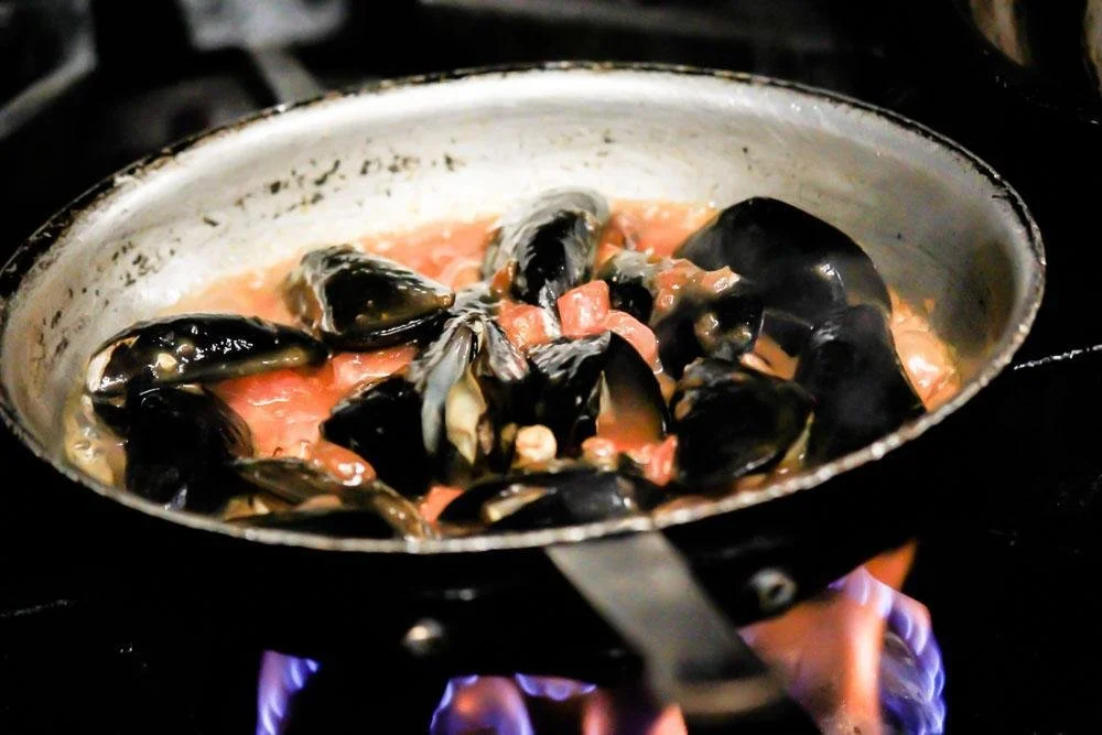 Black mussels cooking in a creamy tomato sauce in a stovetop pan over an open flame.