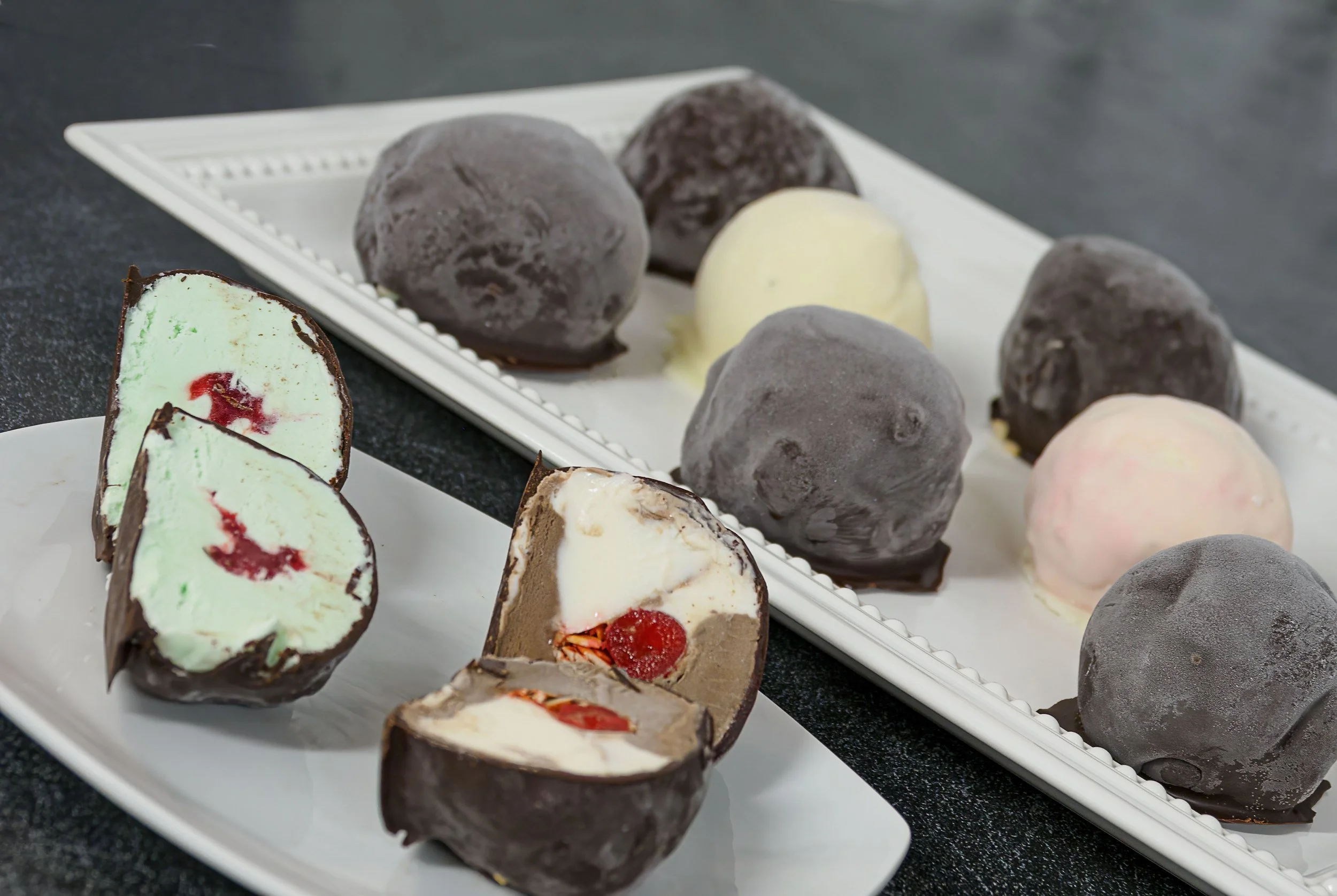 Assorted chocolate-covered ice cream or gelato balls on white trays, with two sliced open to show interior flavors and fillings.
