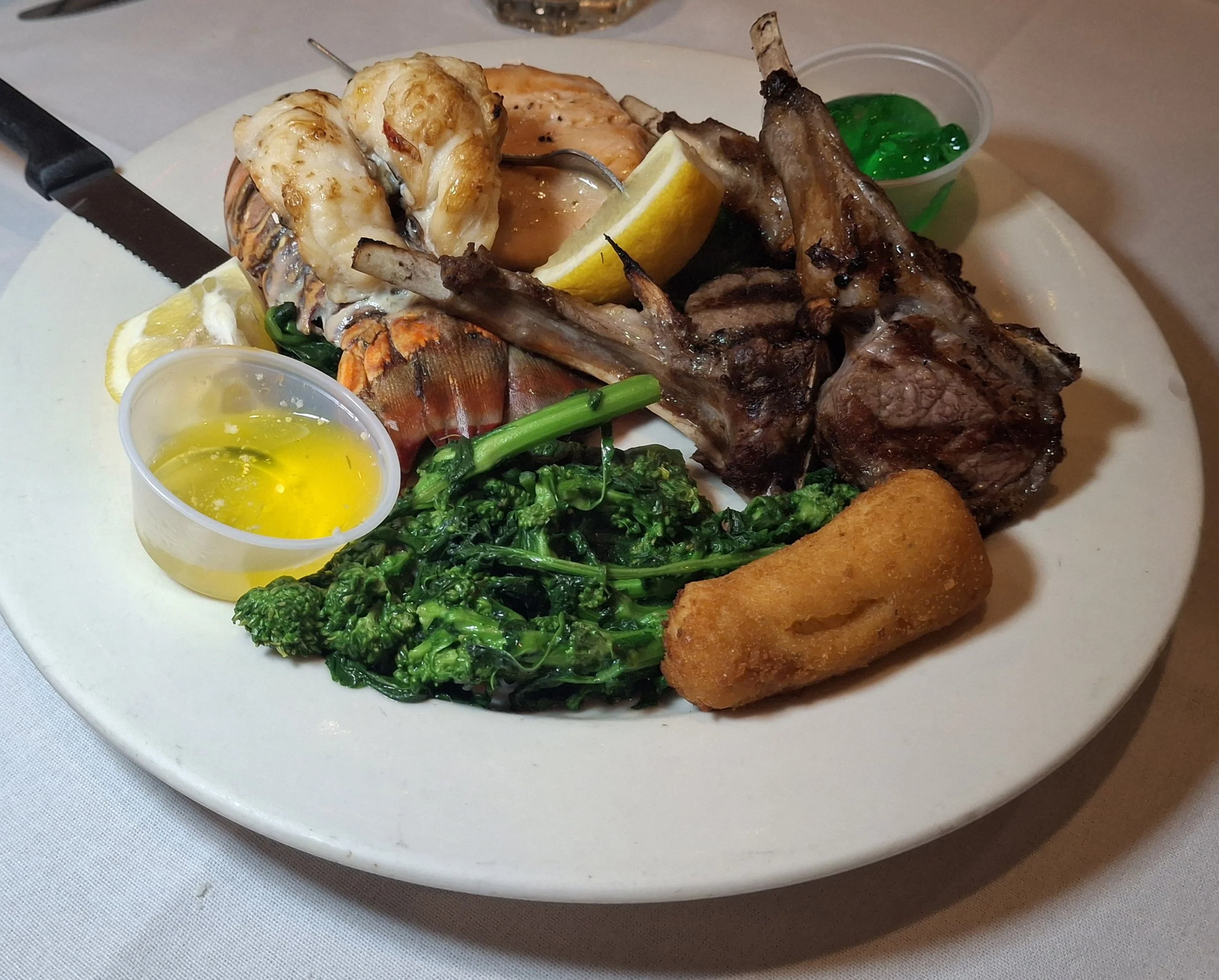 Seafood platter with lobster tail, crab leg, grilled lamb chops, fried croquette, sautéed broccolini, lemon wedges, and small containers of green and yellow sauces.