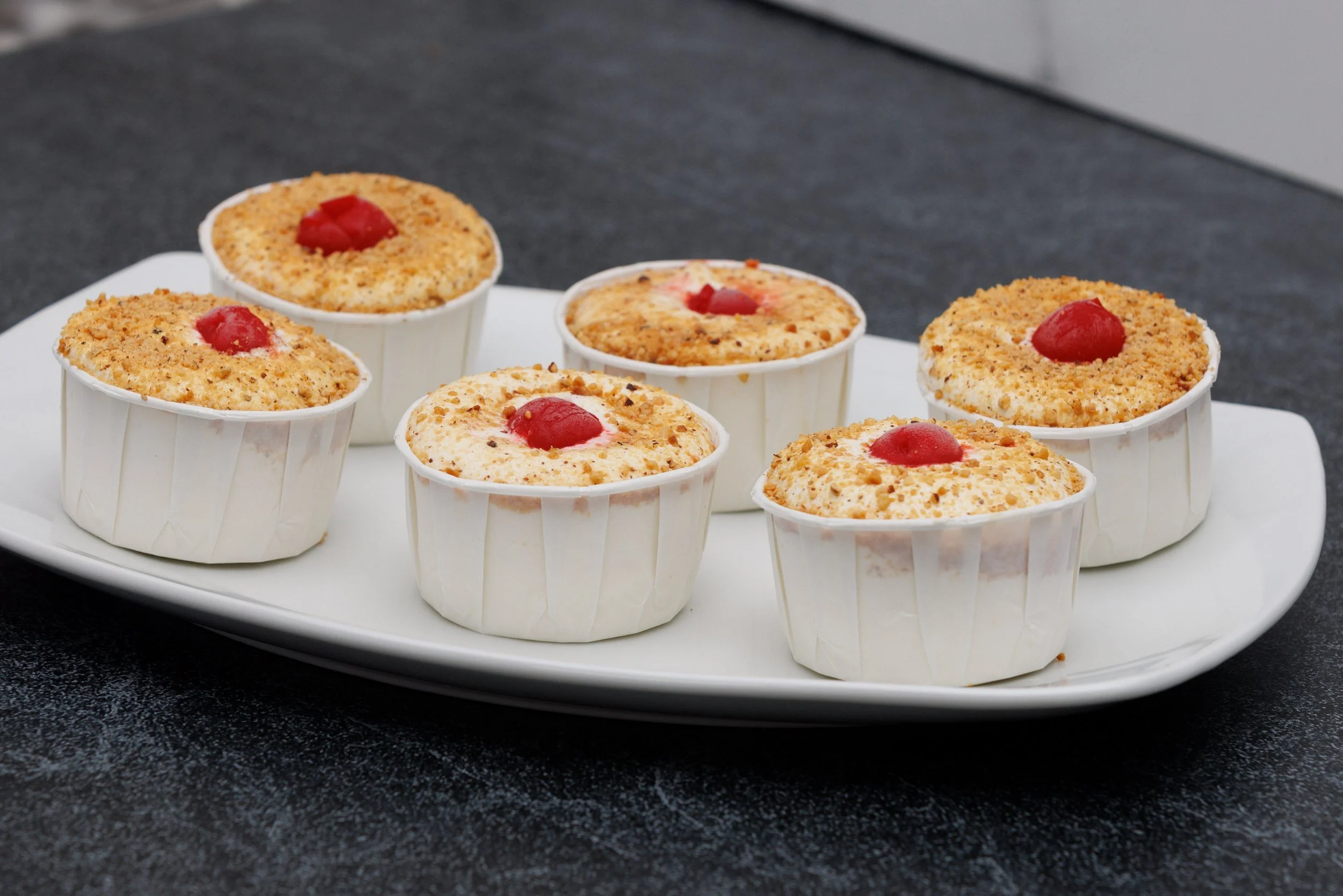 Six individual cheesecakes in white paper cups with red cherry toppings on a white rectangular plate, placed on a black surface.