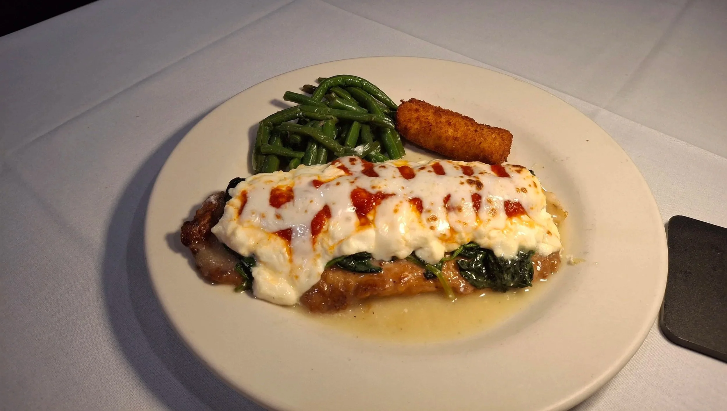 Plate of baked dish topped with melted cheese, side of green beans, and a breaded fried item, on a white tablecloth.
