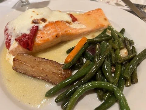 Salmon fillet topped with melted cheese and tomato sauce, served with green beans, carrots, and a piece of pork belly on a white plate.