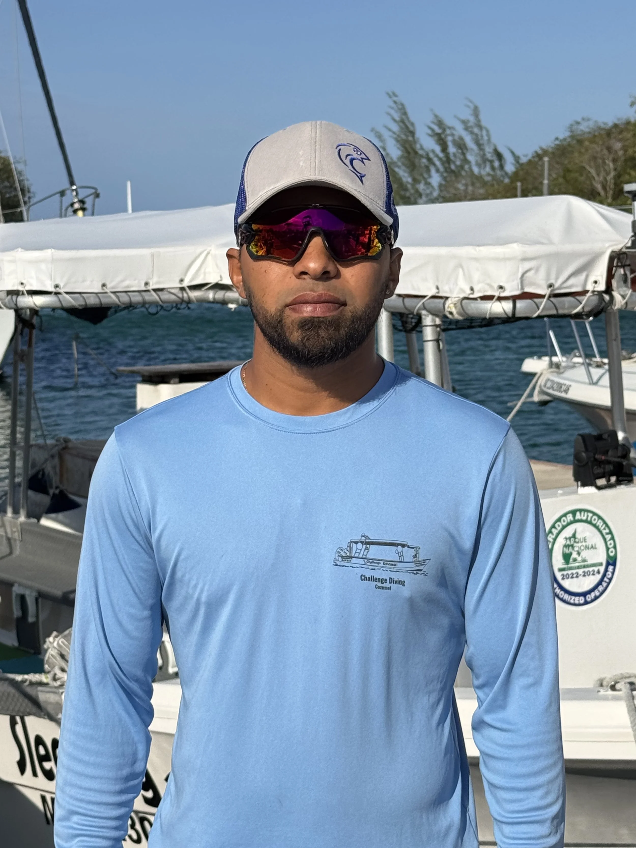 Challenge Diving Dive Master, Jr. in sunglasses and a baseball cap standing on a boat with water and trees in the background.