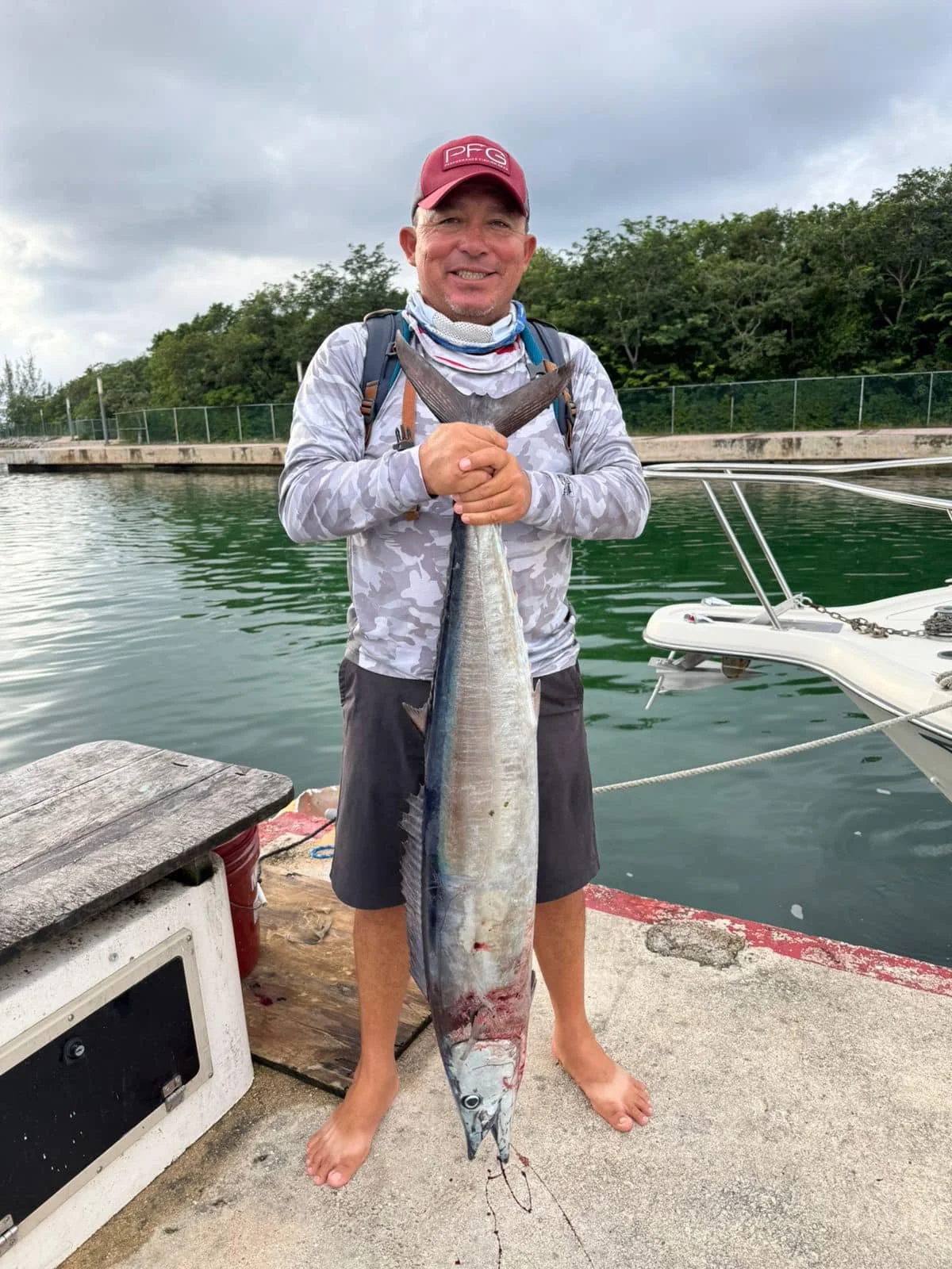 A man wearing a red cap and gray long-sleeve shirt standing on a dock holding a large fish. The man is smiling and has a backpack. The water and a small boat are visible in the background, with trees beyond. The man is barefoot.