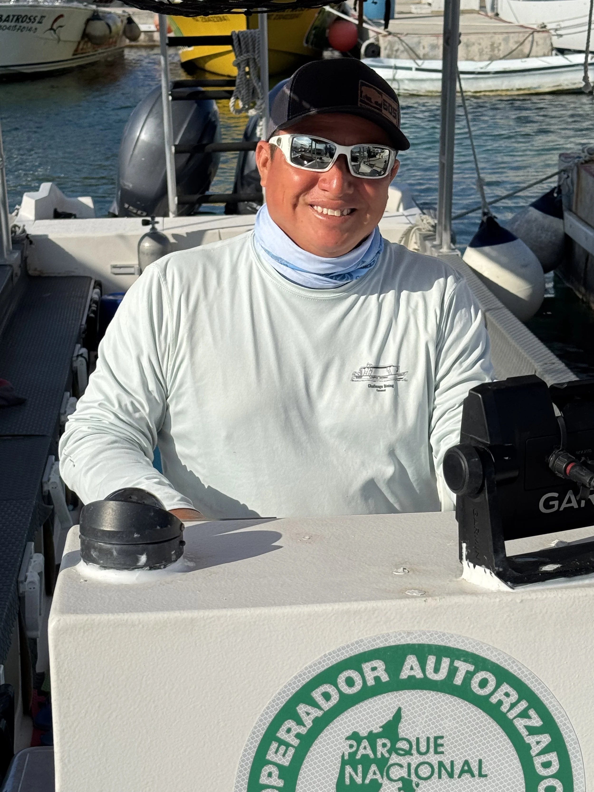 Challenge Diving Baby Shark Captain Ruben Smiling man wearing sunglasses, cap, and white long-sleeve shirt sitting on a boat dock with boats in the background.