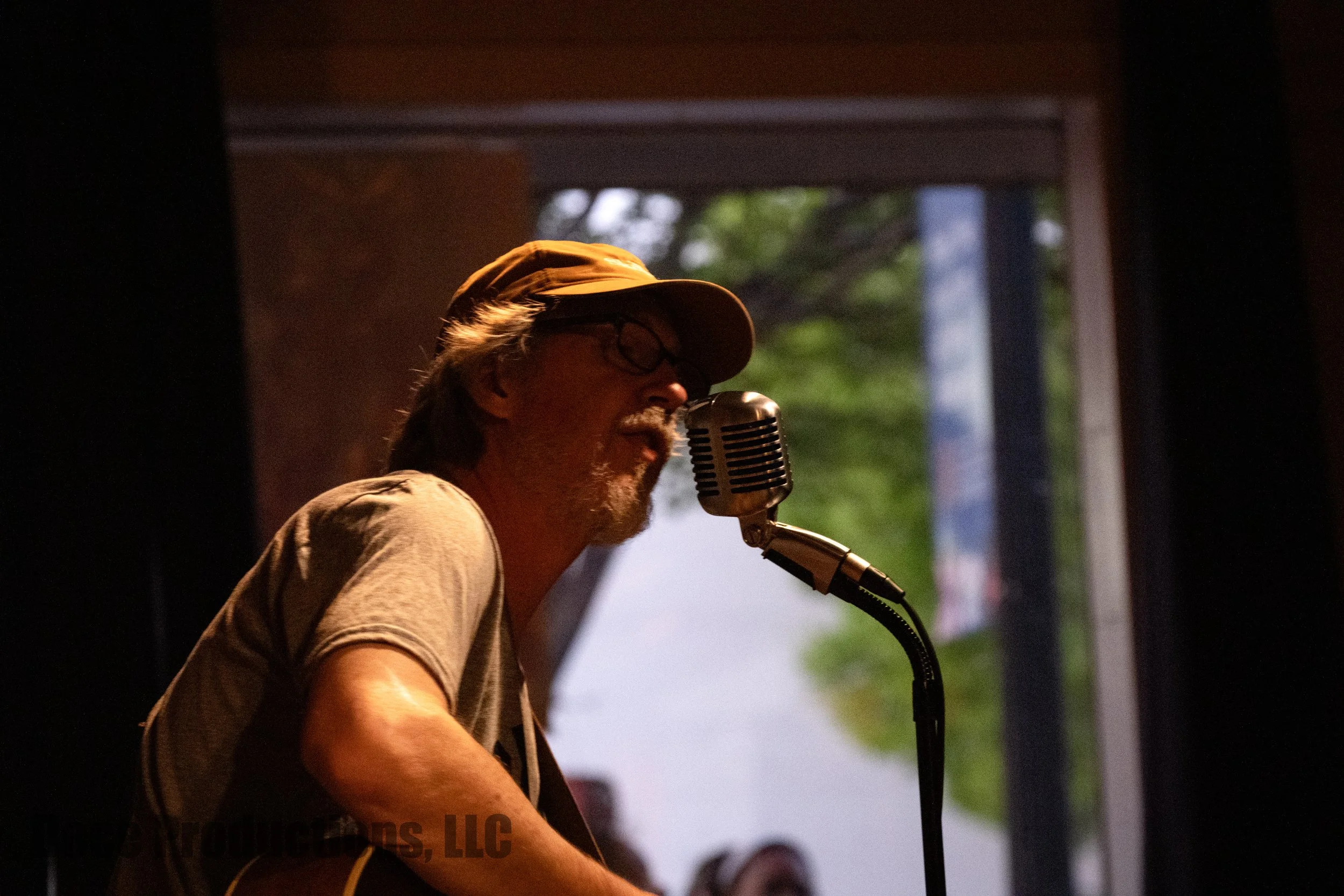 Bearded man wearing a baseball cap and glasses singing into a vintage microphone onstage.