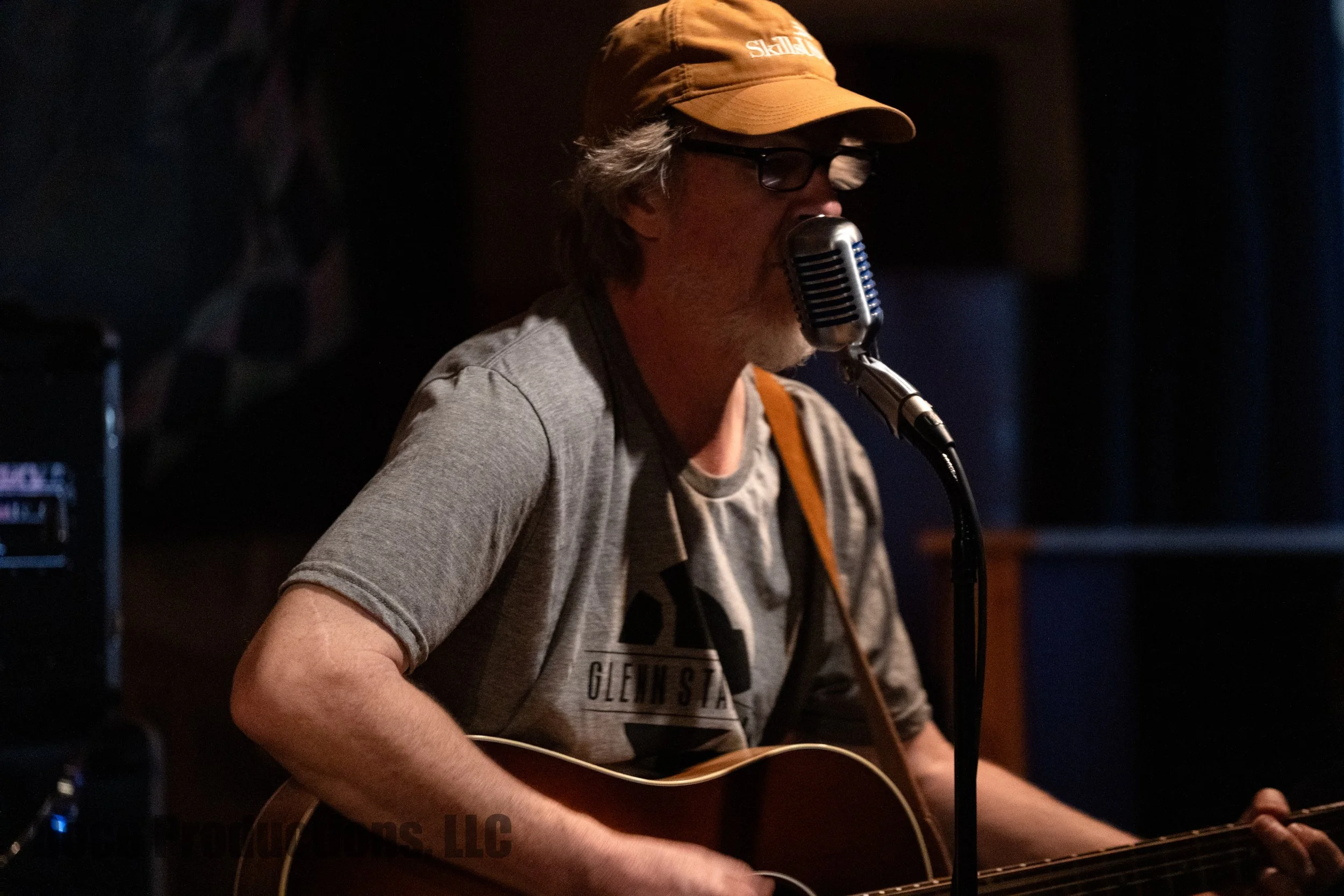 Musician wearing a baseball cap and sunglasses sings into a vintage microphone while playing acoustic guitar onstage.