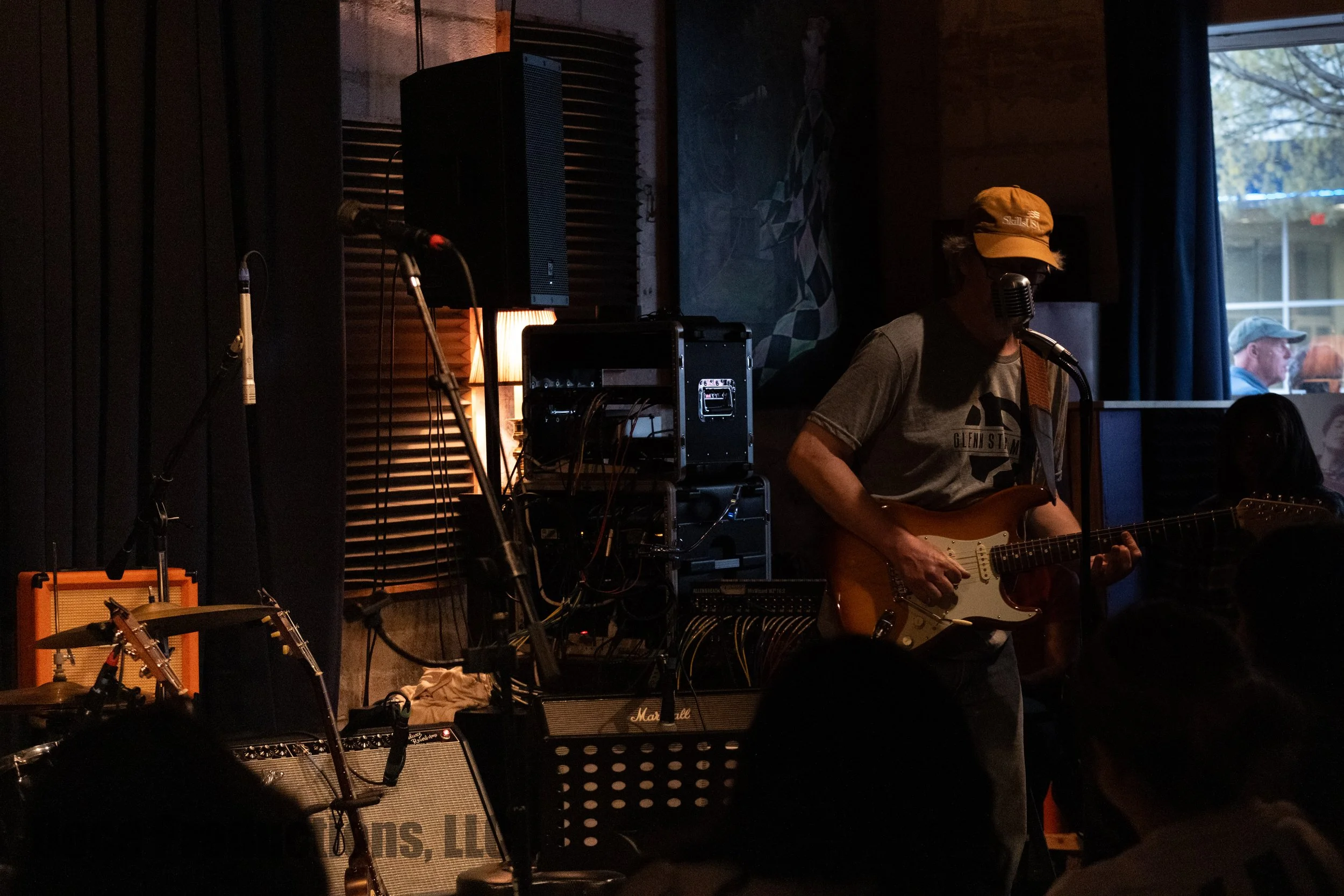 Musician playing electric guitar and singing into a microphone at an indoor live performance surrounded by amplifiers and audio equipment