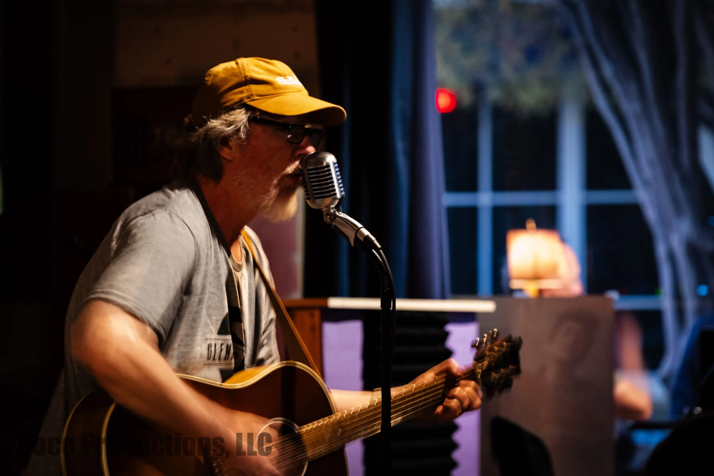 Musician wearing a yellow cap and glasses sings into a vintage microphone while playing acoustic guitar on stage.