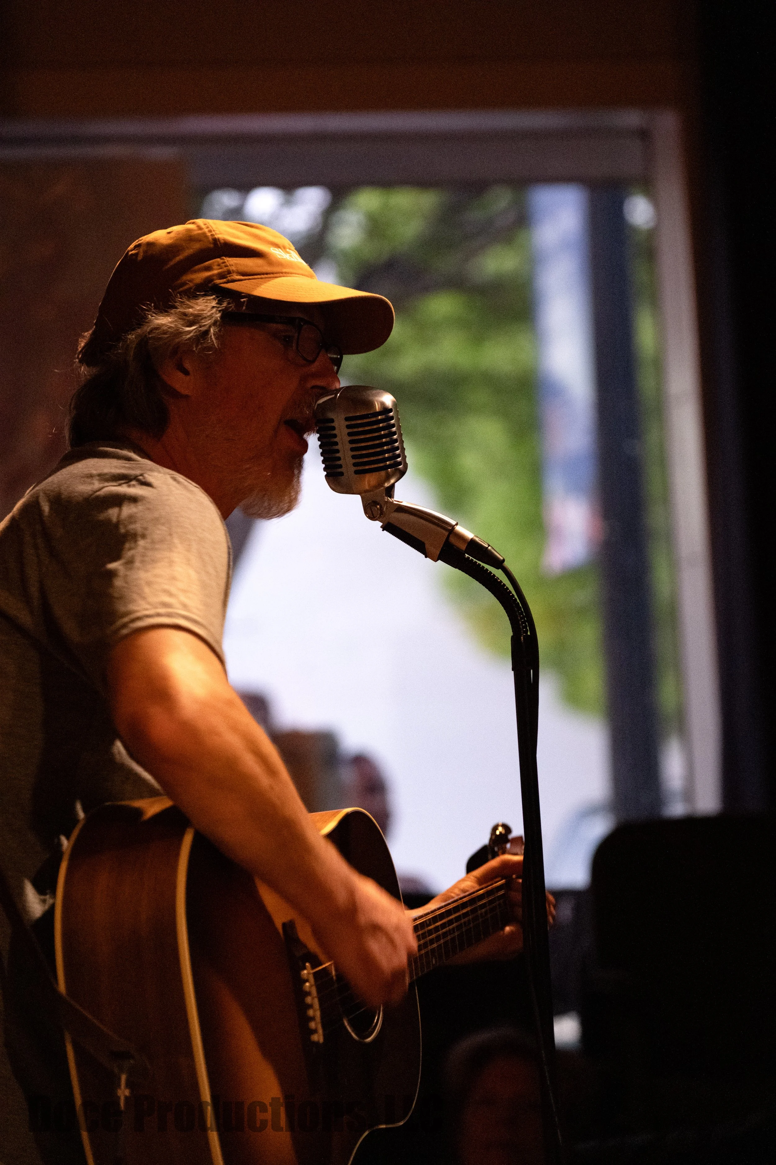 Musician in an orange cap playing an acoustic guitar and singing into a vintage microphone on stage