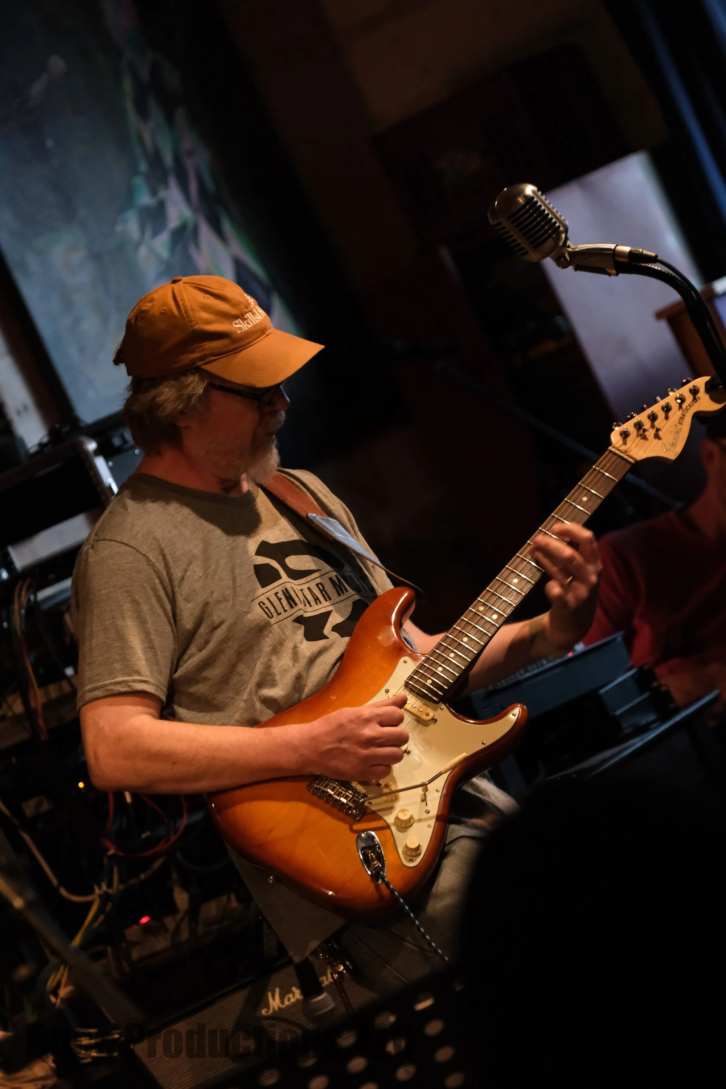 Musician in an orange cap playing an electric guitar beside a vintage microphone onstage