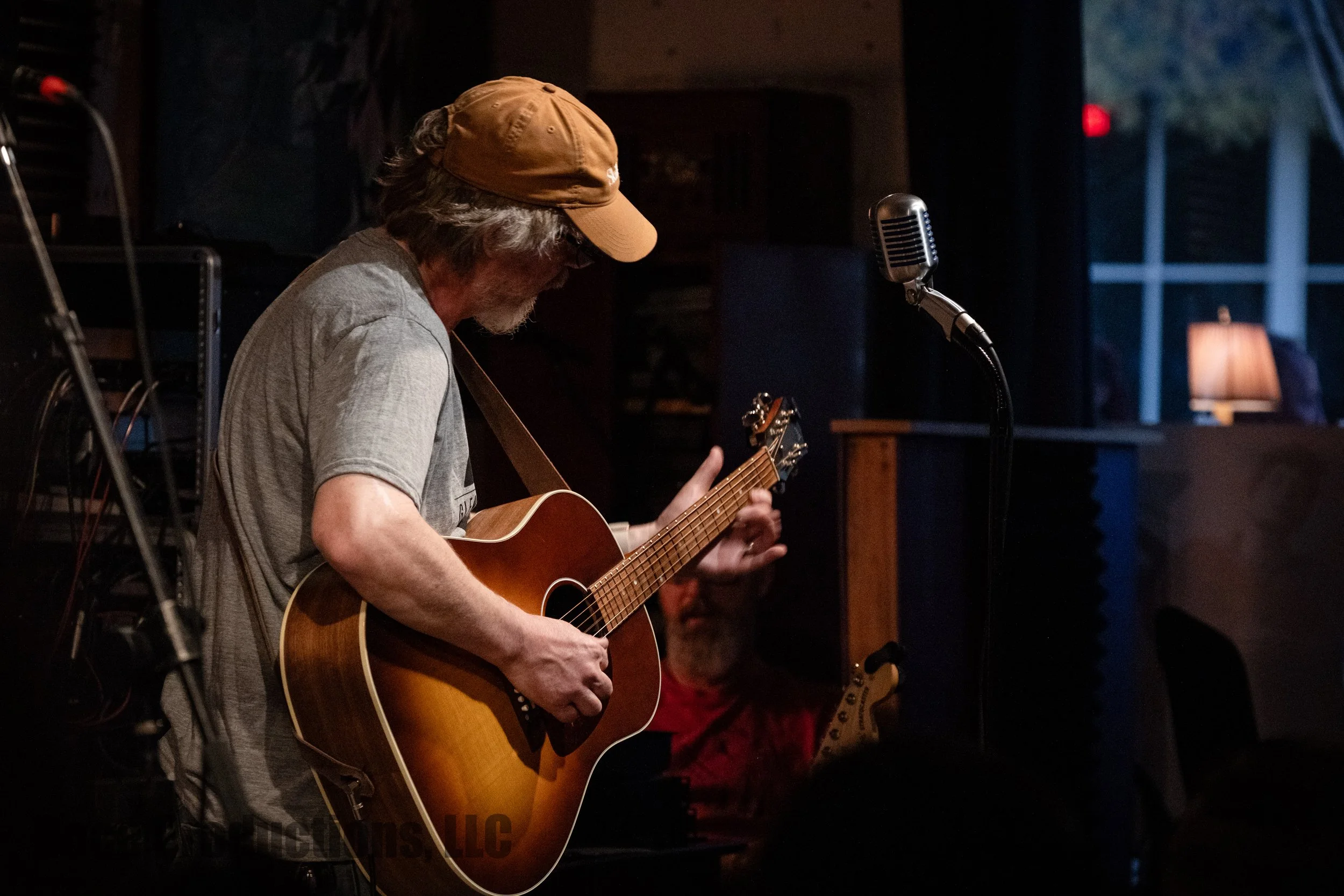 Musician in a baseball cap playing an acoustic guitar beside a vintage microphone on a dimly lit stage